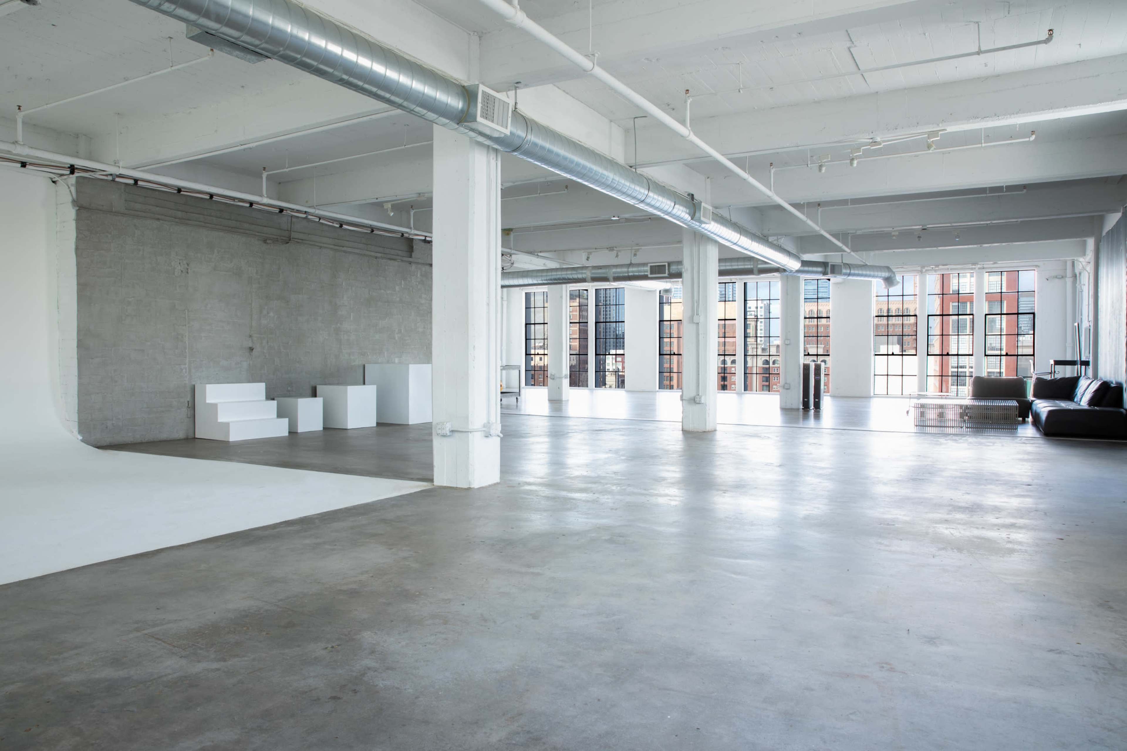 The image shows a spacious, industrial-style studio with concrete flooring, large windows, and ample natural light.