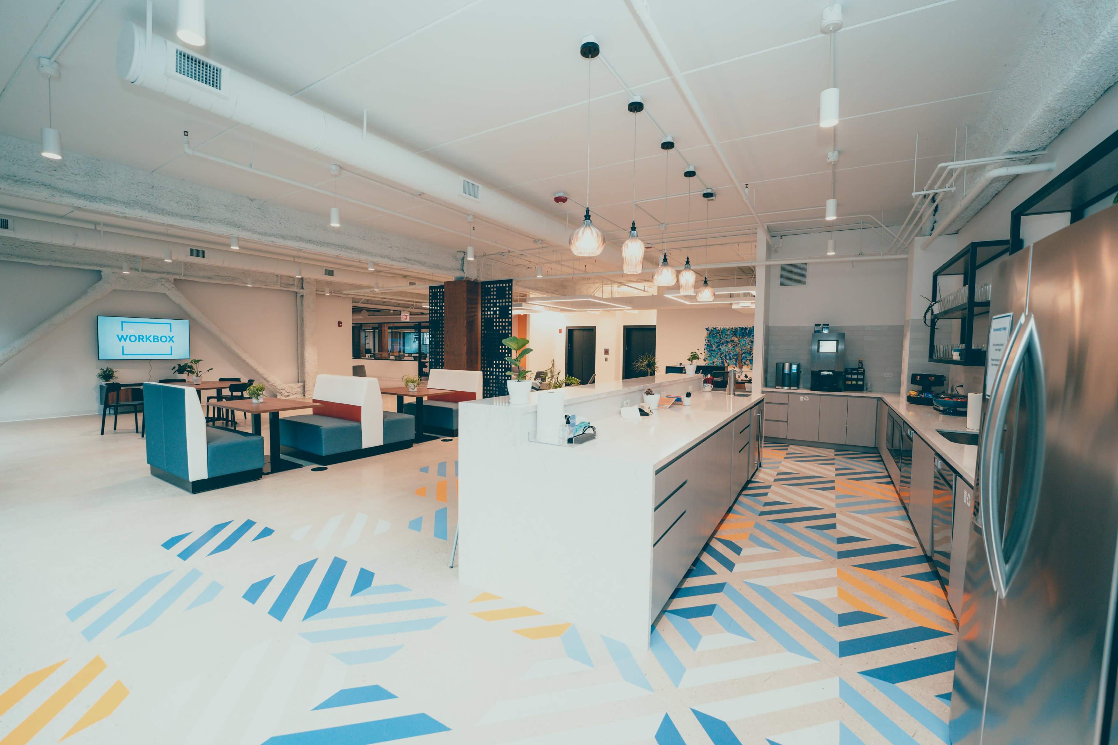 A modern office kitchen area with a large central island, colorful geometric-patterned flooring, and seating arrangements in a spacious environment.