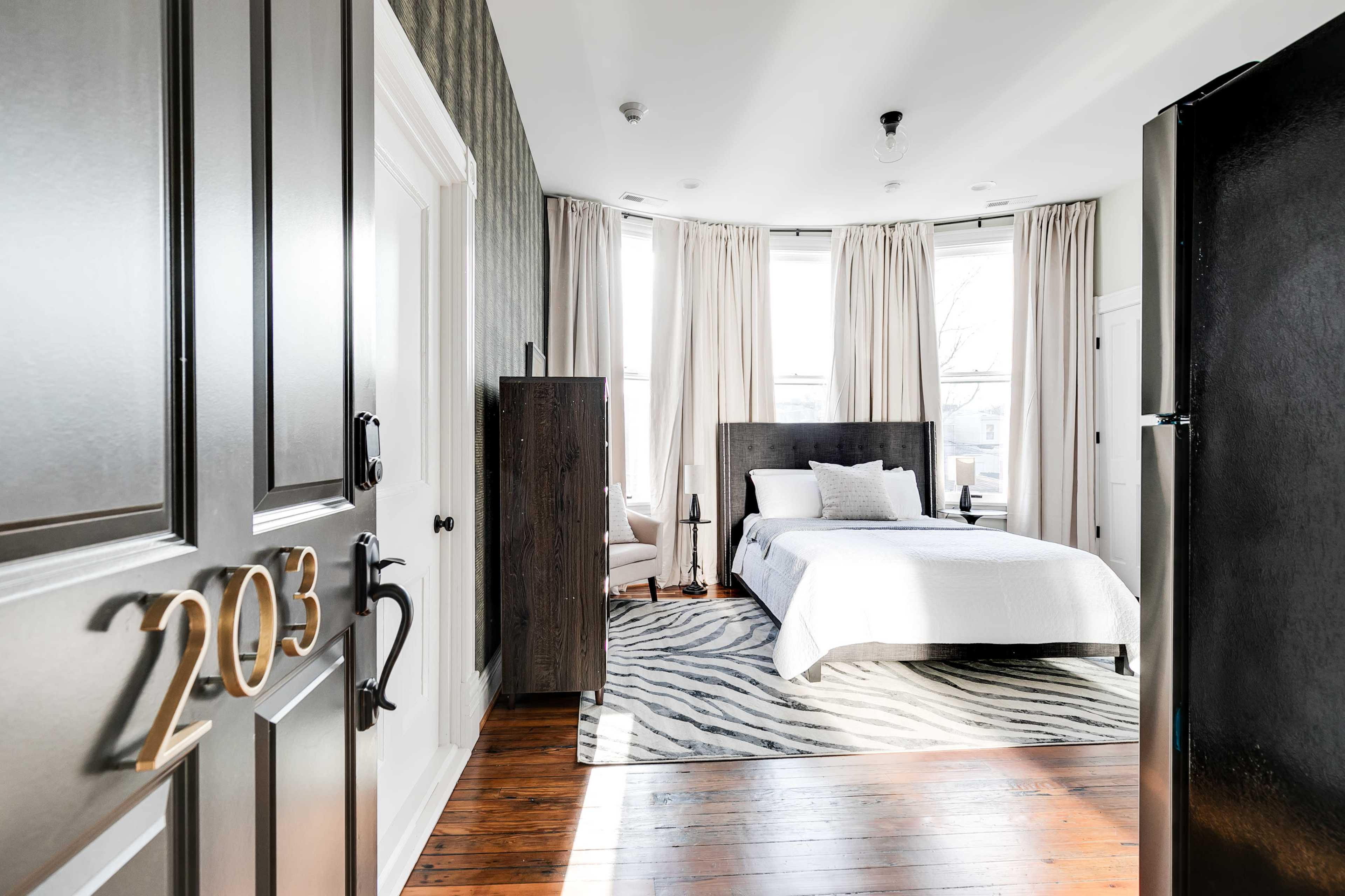 A modern hotel room with a bed, large windows, and a door marked "203" in a corridor.