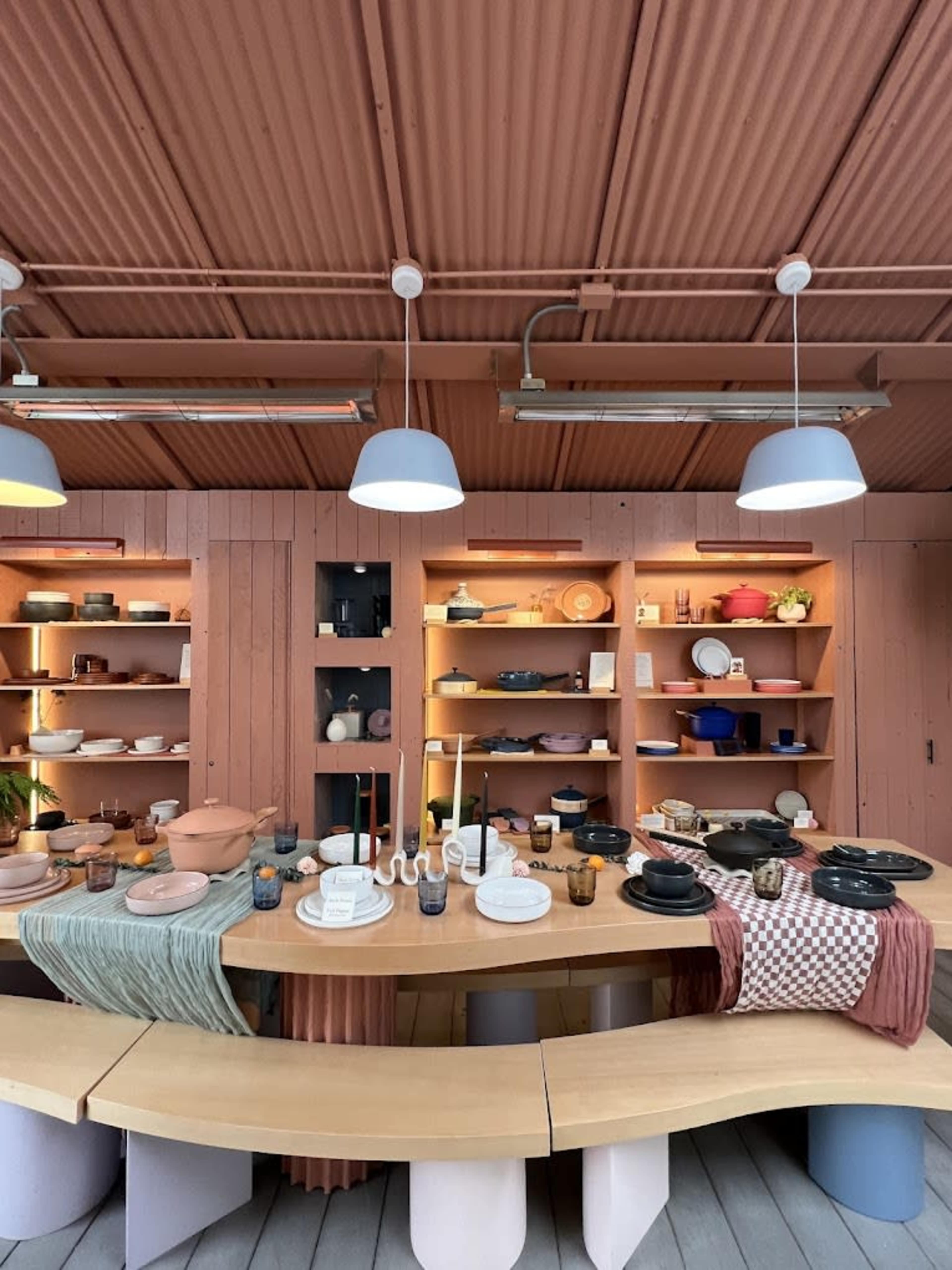 The image shows a kitchen space with wood-paneled walls, shelves filled with various kitchenware, and a large, curved wooden table set with plates and glasses.
