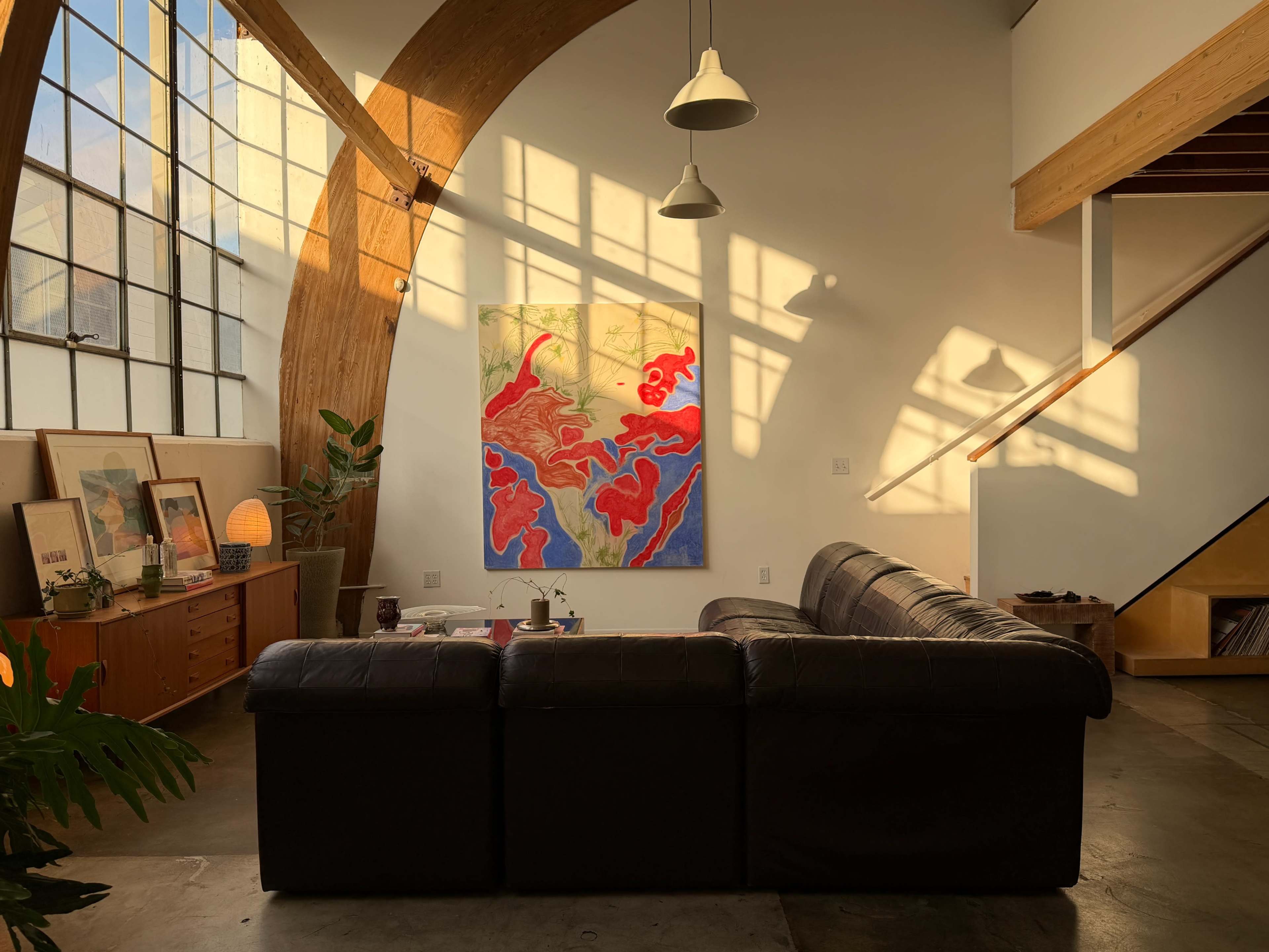 A modern living room features a large abstract painting, a leather couch, and sunlight streaming through tall windows, casting shadows on the walls.