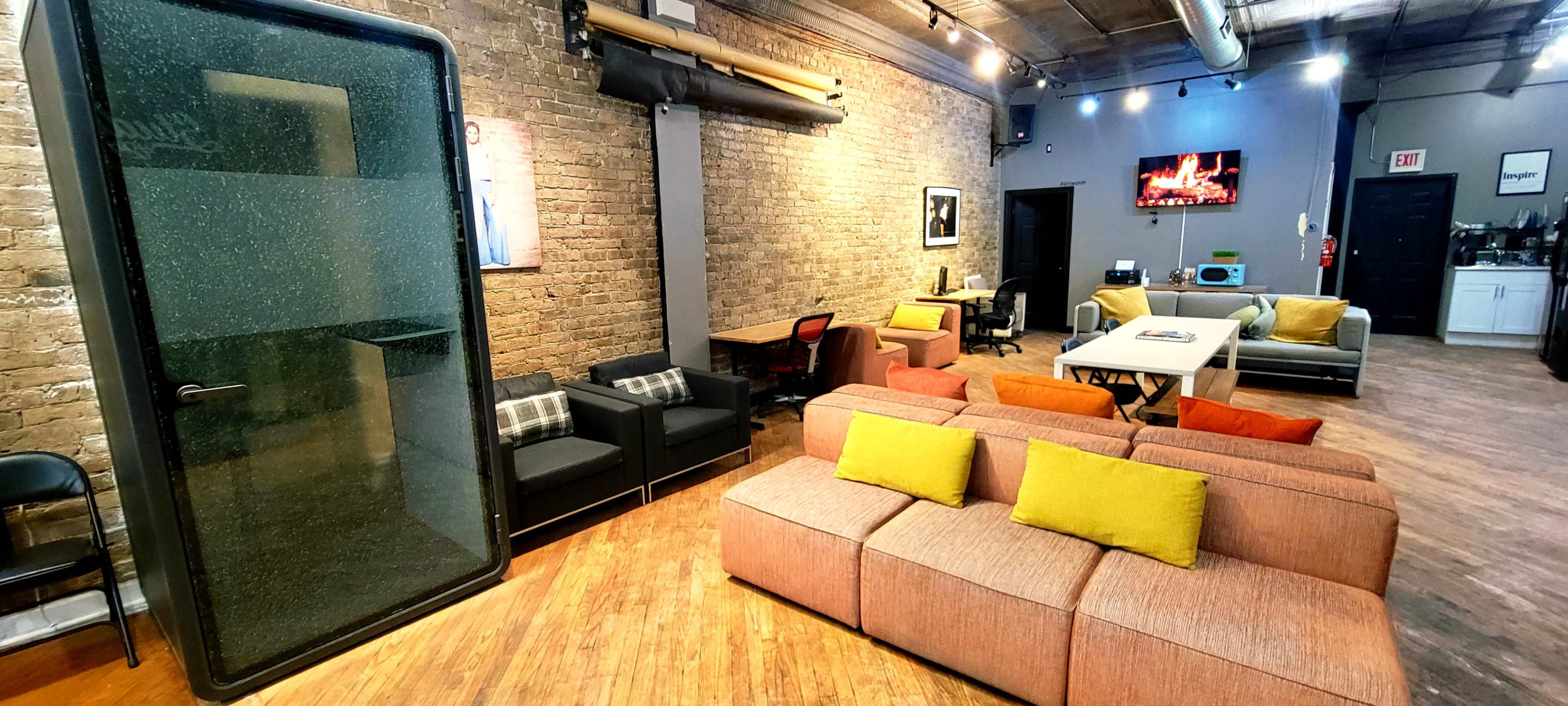 The image shows a modern interior of a communal workspace featuring a lounge area with various seating options, a partitioned glass meeting room, and a kitchenette in the background.