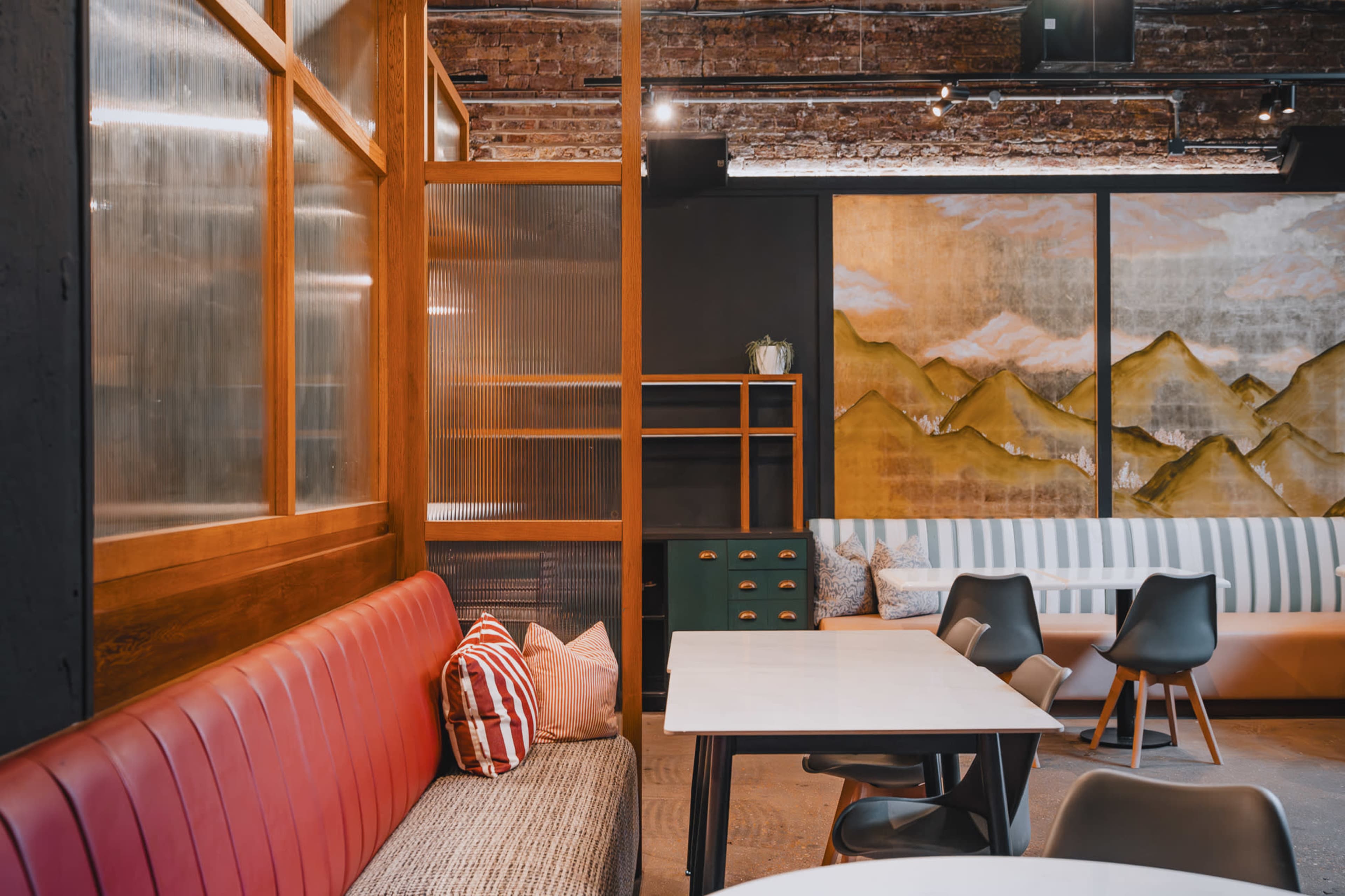 The image shows an interior space of a café featuring a mix of wooden elements, a red upholstered bench, and a striped sofa against a mural of mountains.