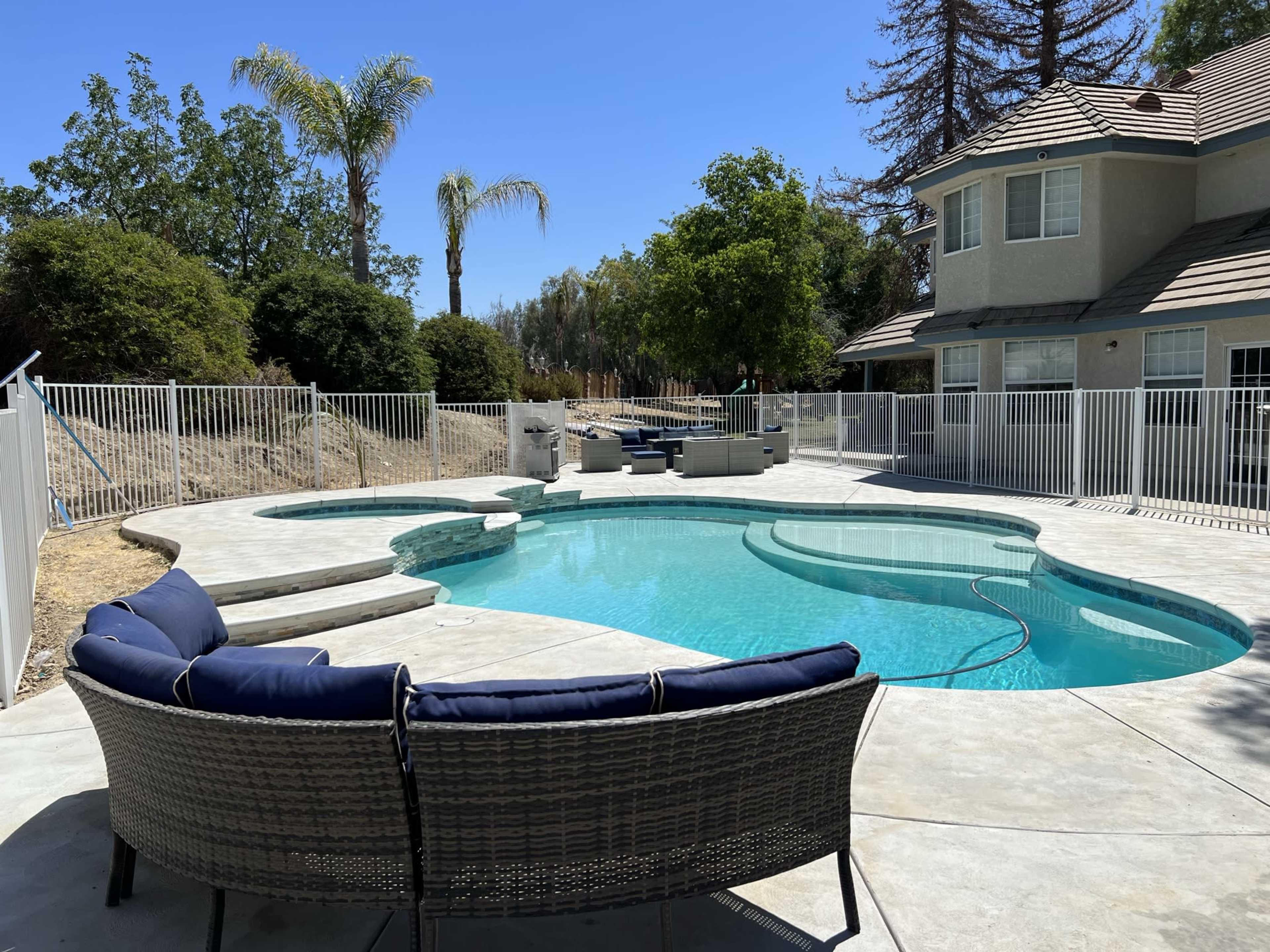 The image shows a curved swimming pool surrounded by a concrete patio and a white fence, with outdoor seating and palm trees in the background.