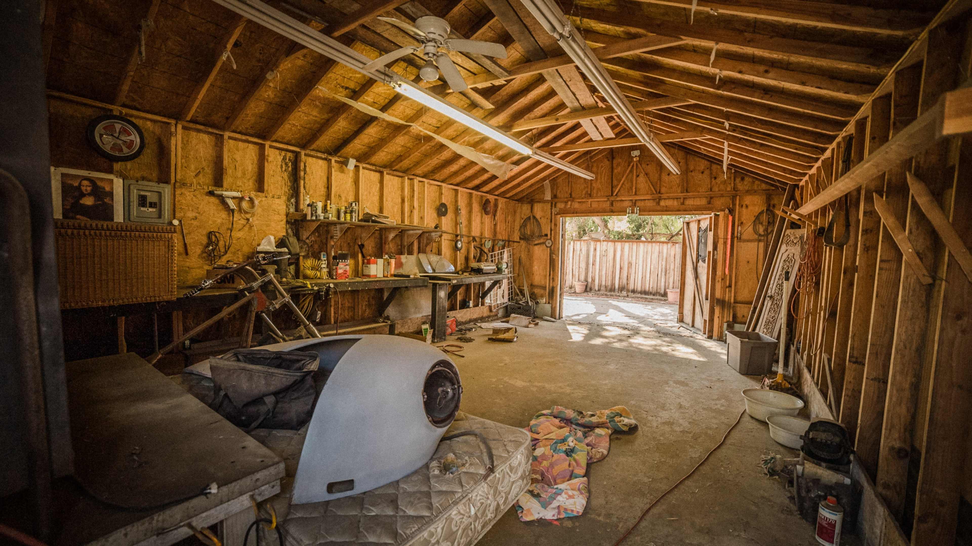The interior of a wooden garage with unfinished walls, featuring a workbench, tools, and an open door leading to an outdoor area.