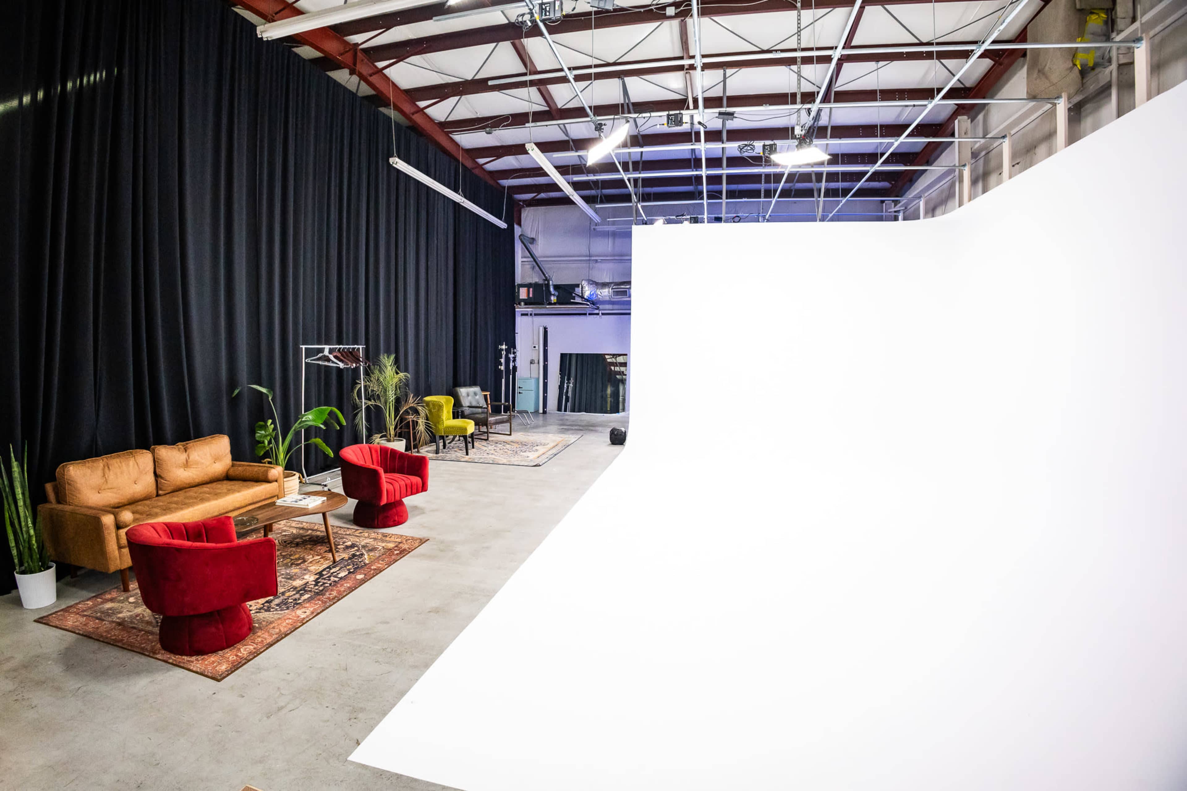 The image shows a spacious studio with a white backdrop, a brown sofa, and red and yellow chairs arranged on a concrete floor, alongside green plants and rugs.