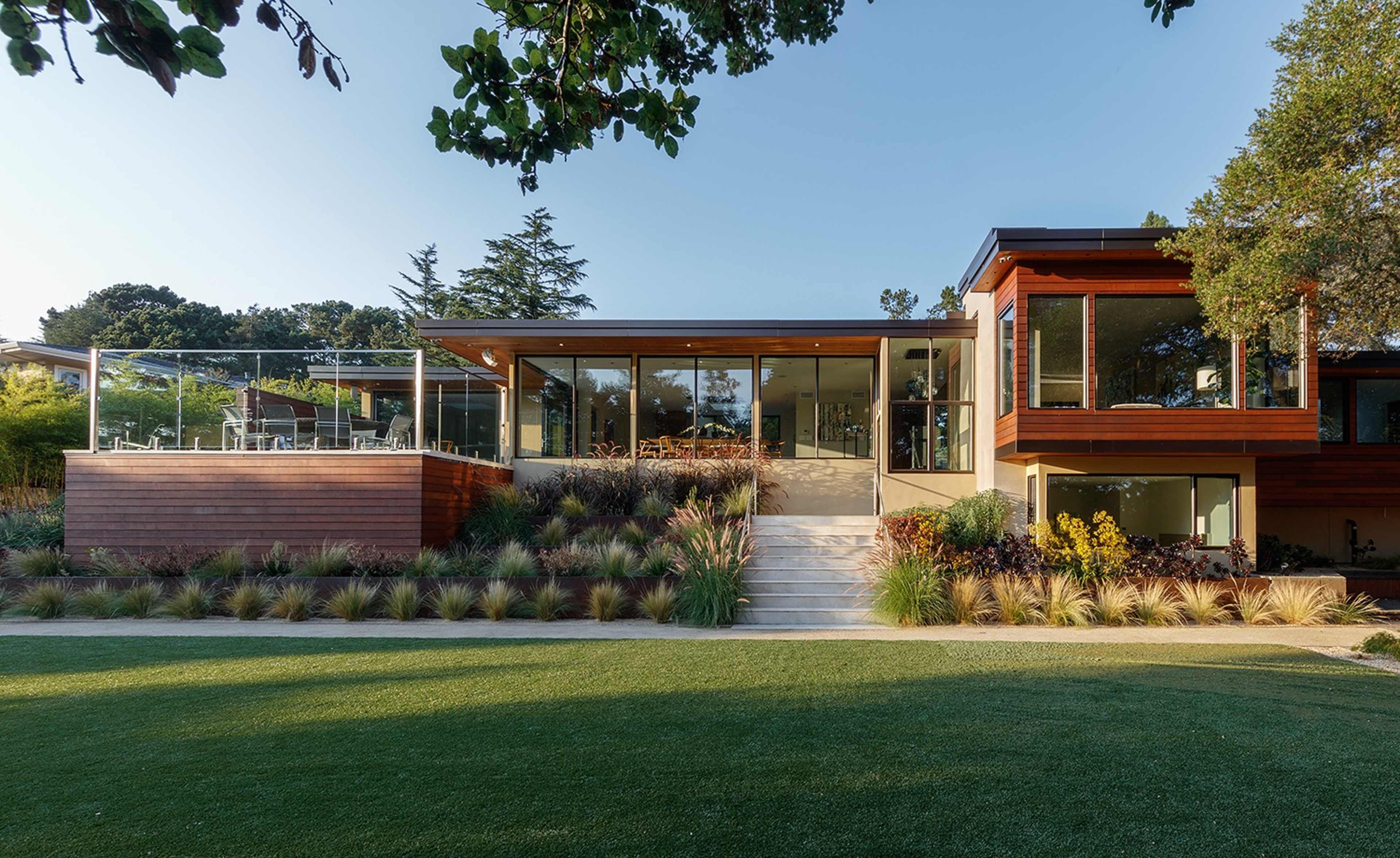 The image showcases a modern house with large glass windows surrounded by landscaped greenery and a patio area.