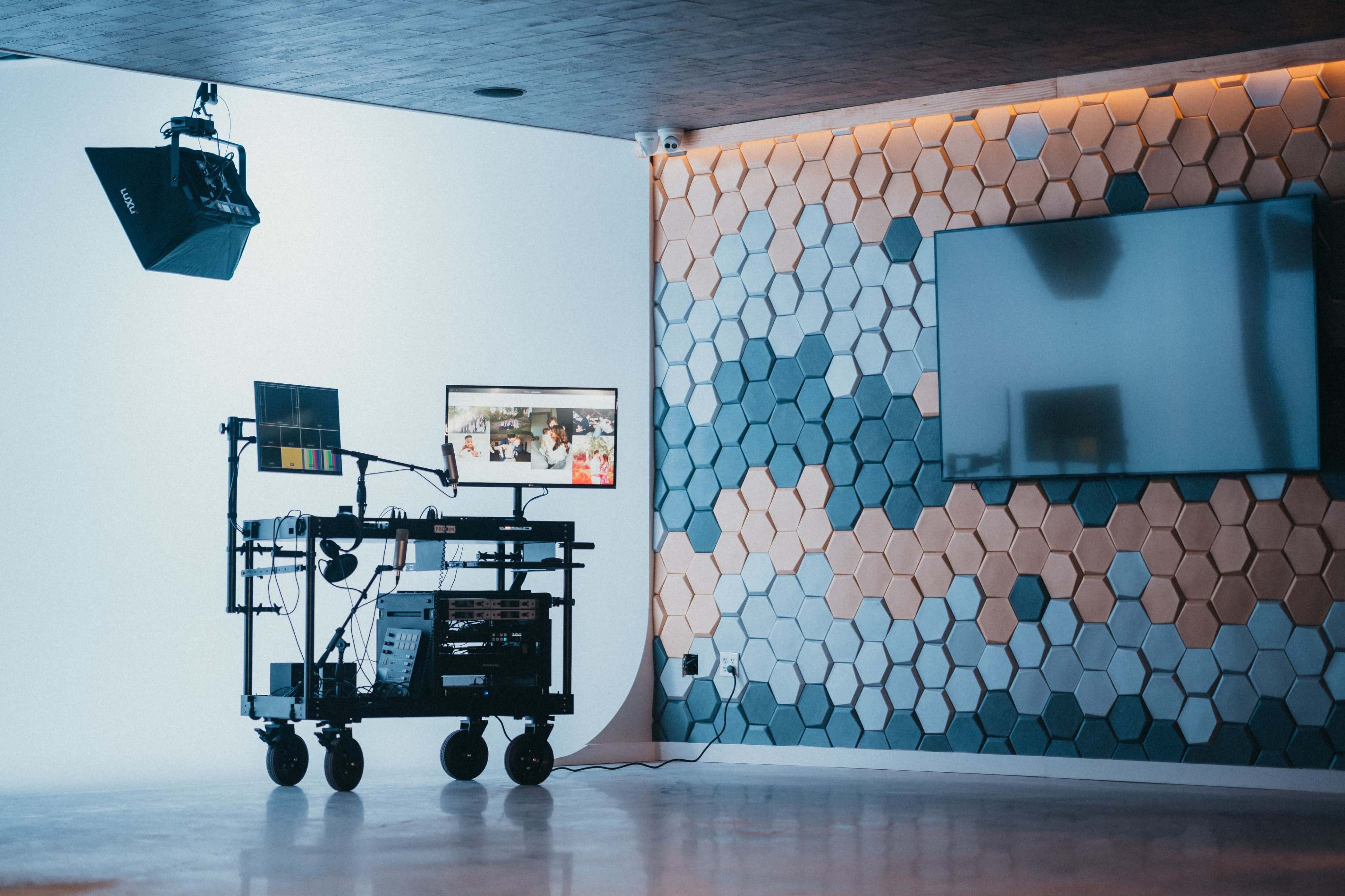 A video production setup featuring a cart with equipment, a mounted monitor, and a large light fixture in a room with a hexagonal patterned wall.