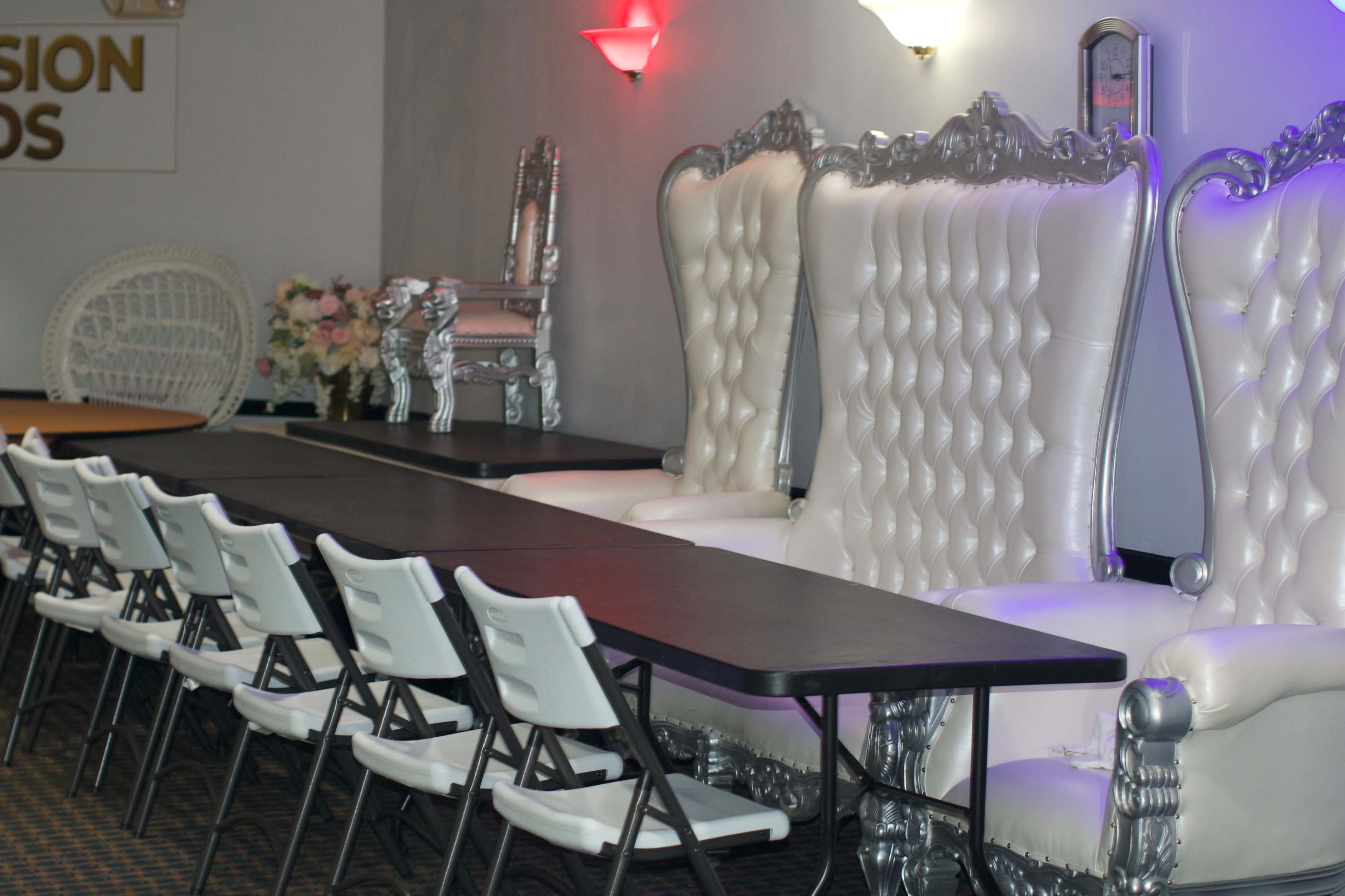 The image shows a room with two large, ornate white chairs at the back and a row of folding chairs in front of a long rectangular table.