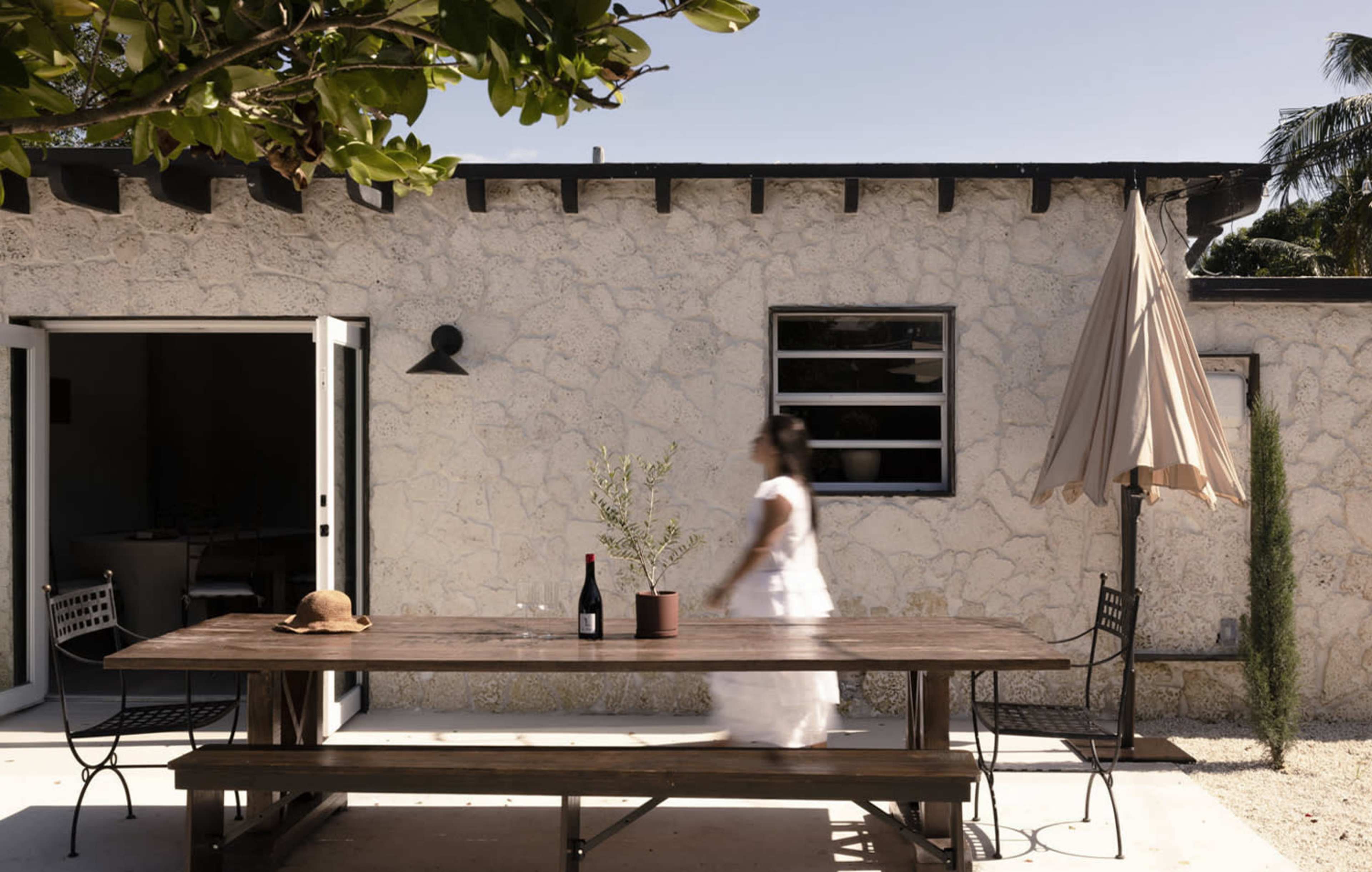 A woman walks past a wooden table set with a wine bottle and a potted plant, beside a stone wall and an umbrella.