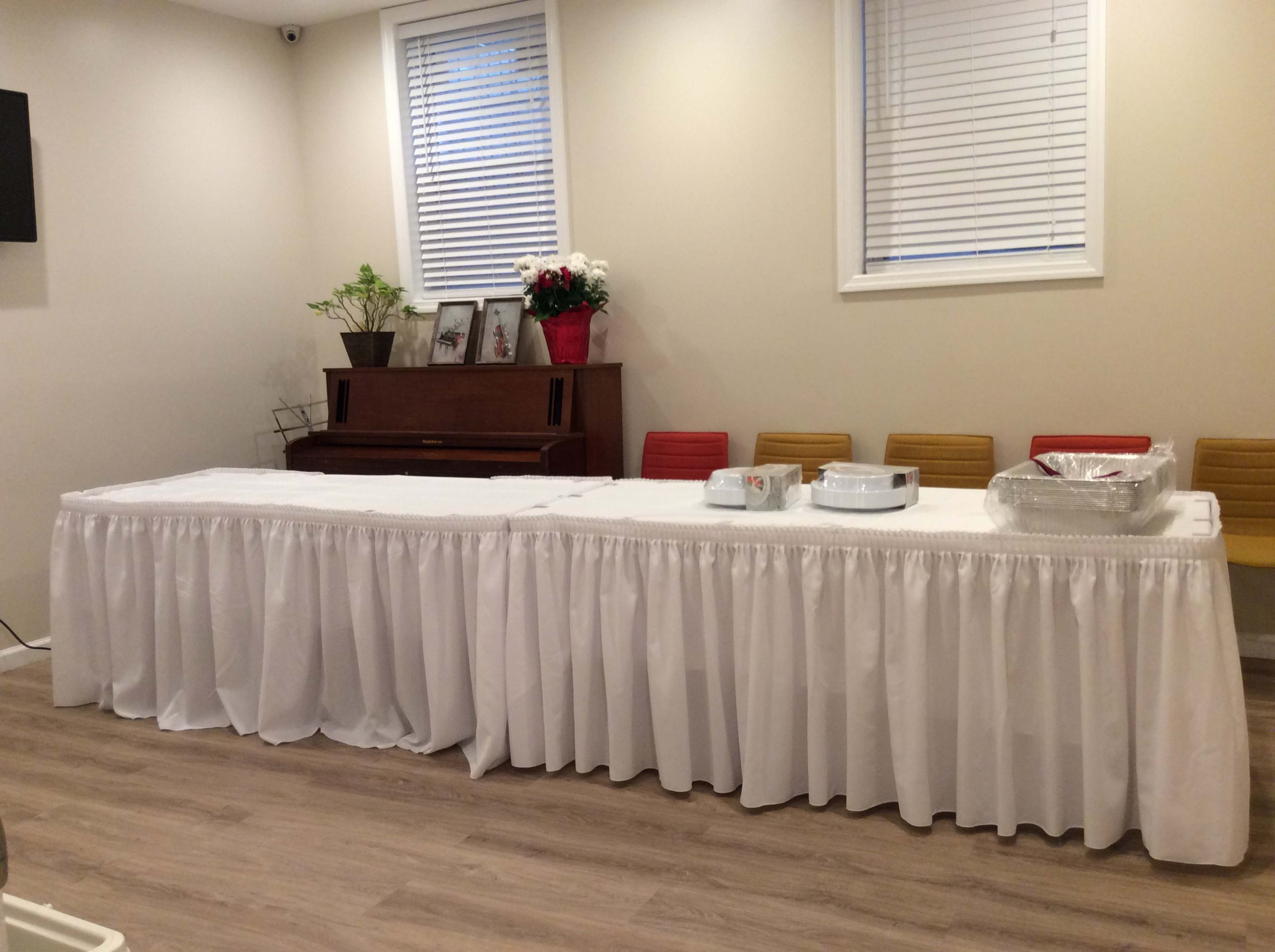 A long table covered with a white tablecloth is set up in a room with a piano and several chairs along the wall.