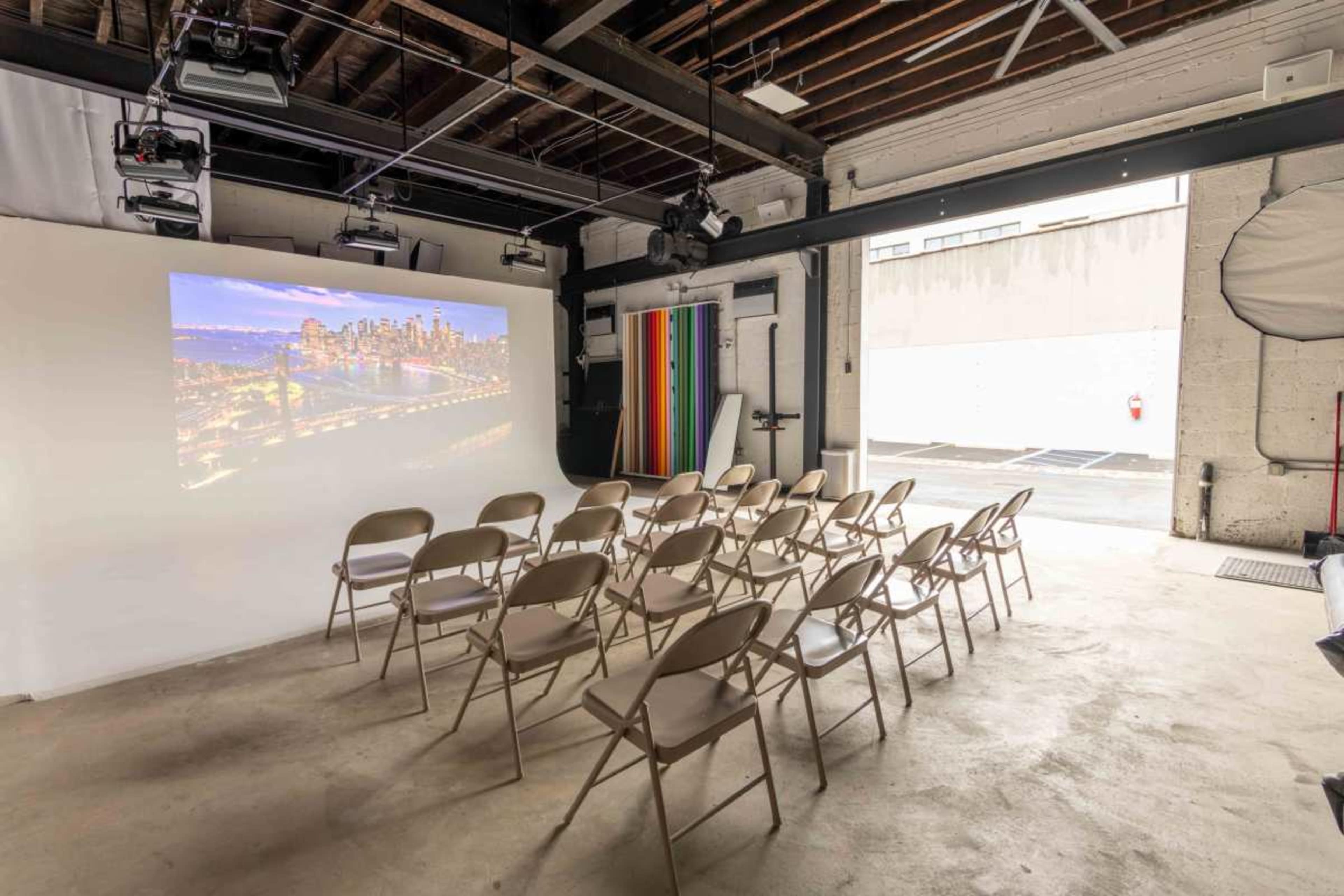 The image shows a minimalist indoor space with rows of folding chairs facing a large projection screen displaying a city skyline, accompanied by colored panels on the side.