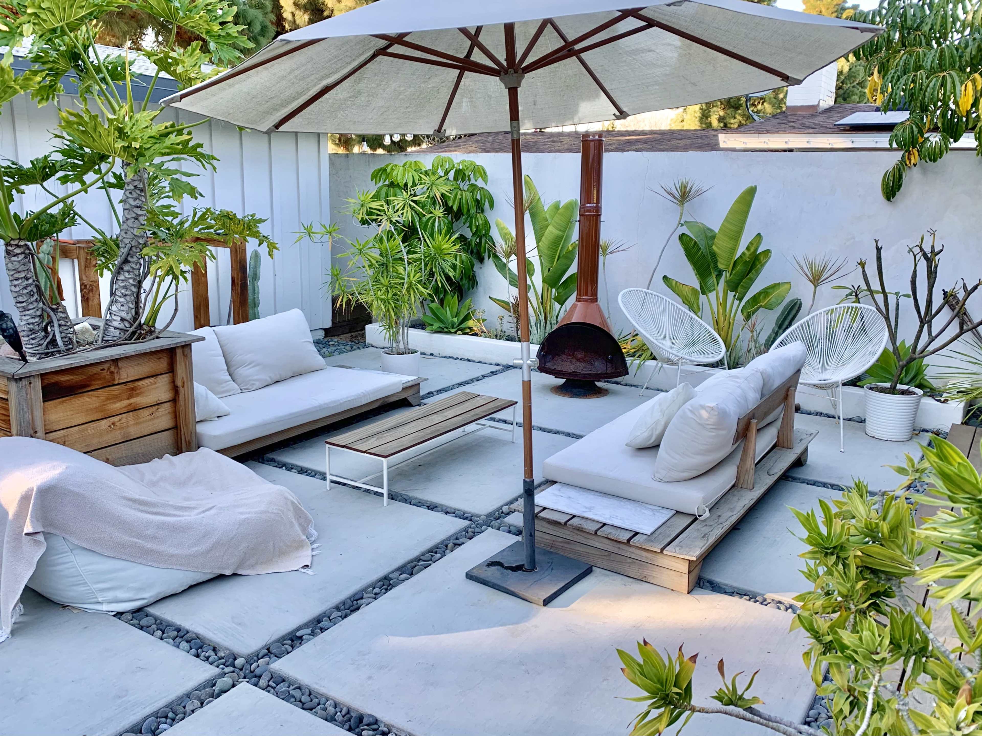 A patio area with two seating arrangements under an umbrella, surrounded by potted plants and a small outdoor fireplace.