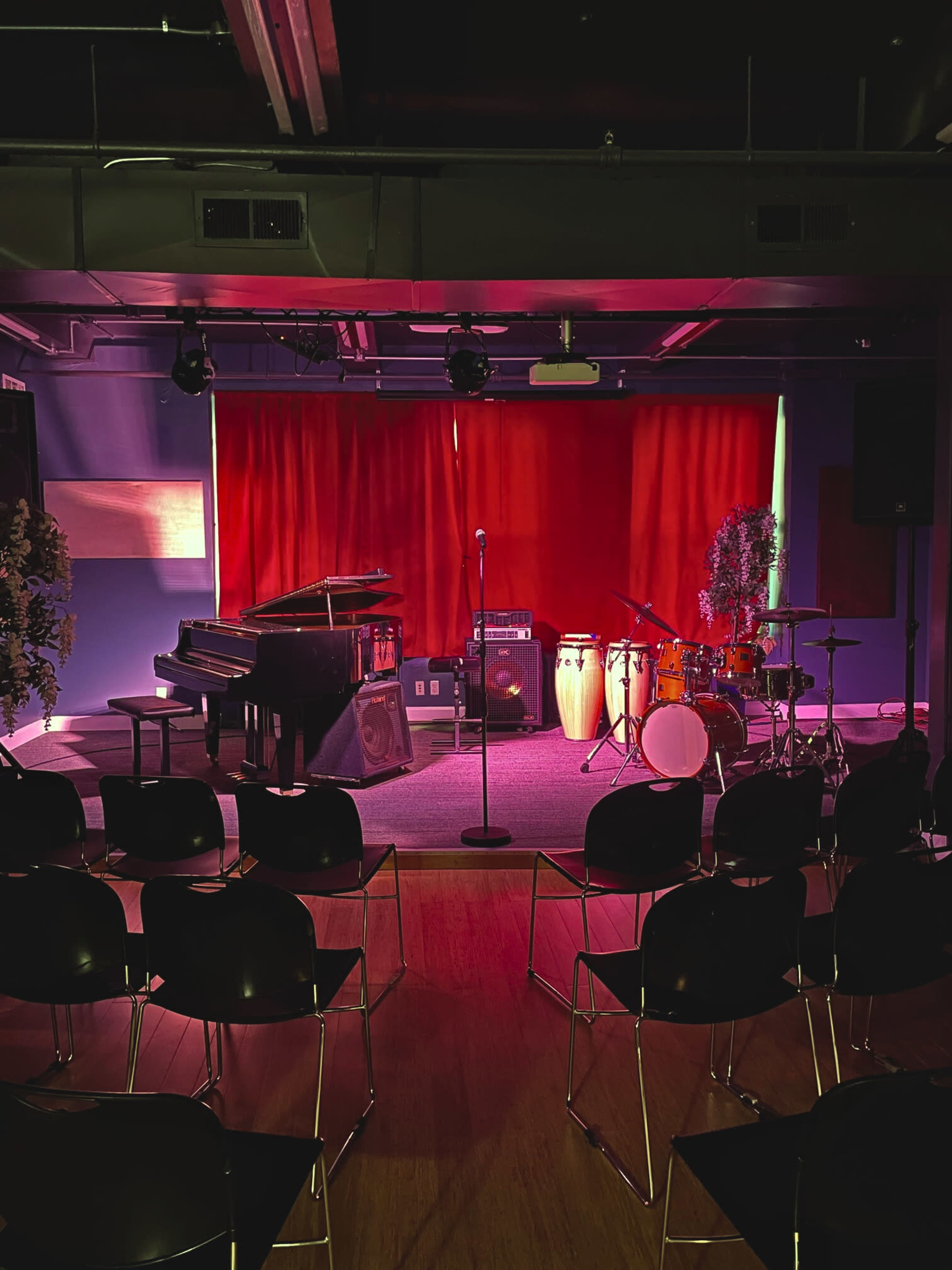 The image shows a stage set up for a performance, featuring a piano, conga drums, a microphone stand, and seating arranged in front of a red curtain.
