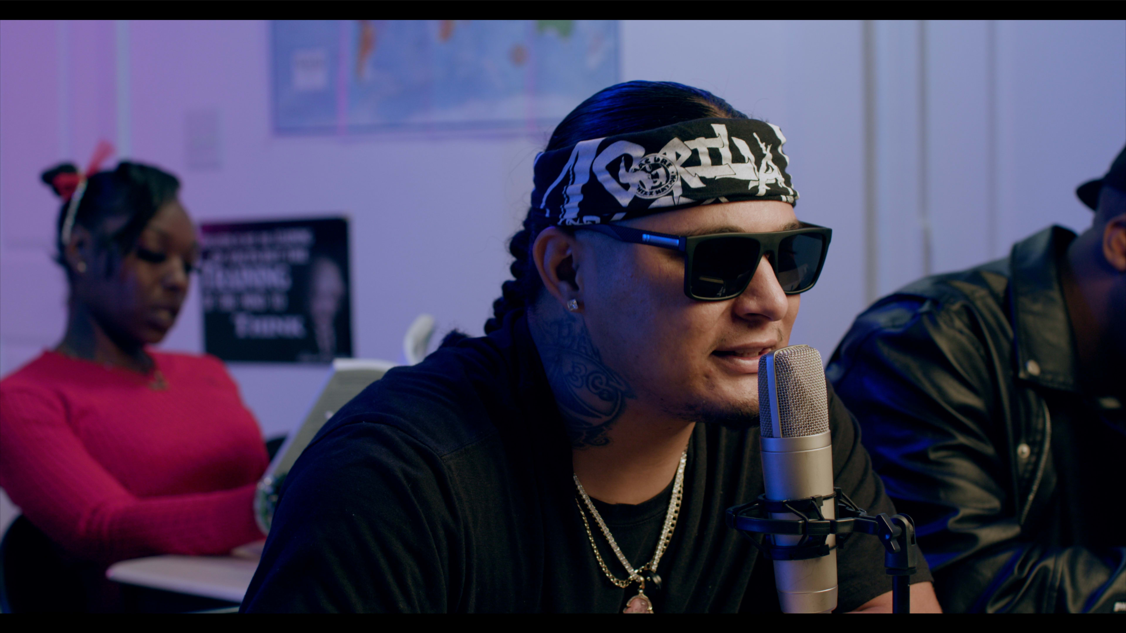 A man wearing sunglasses and a bandana speaks into a microphone, while a woman in a pink sweater sits behind him, focused on her work.