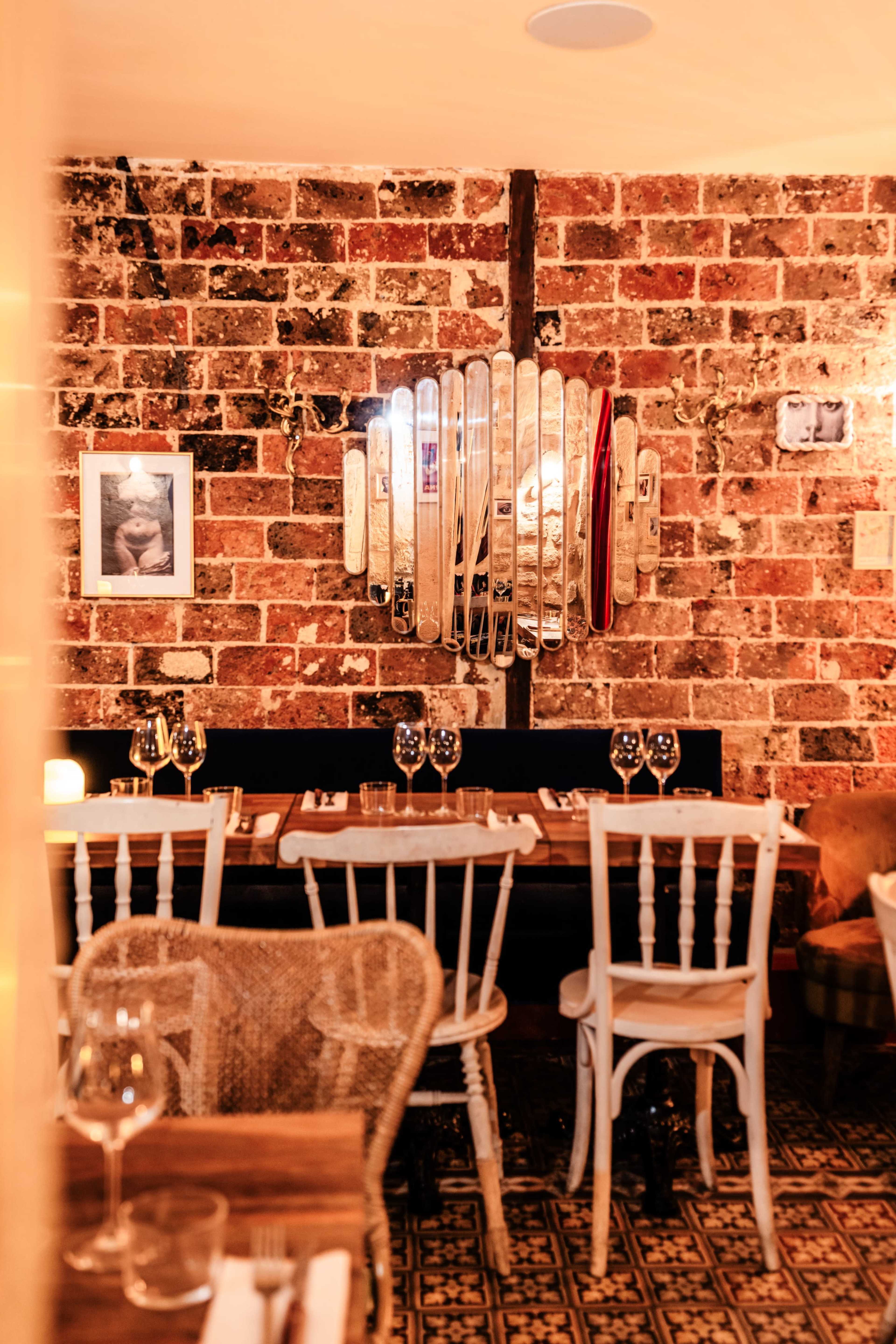A rustic restaurant features a brick wall adorned with a decorative mirror and framed photographs, alongside wooden tables set for dining.