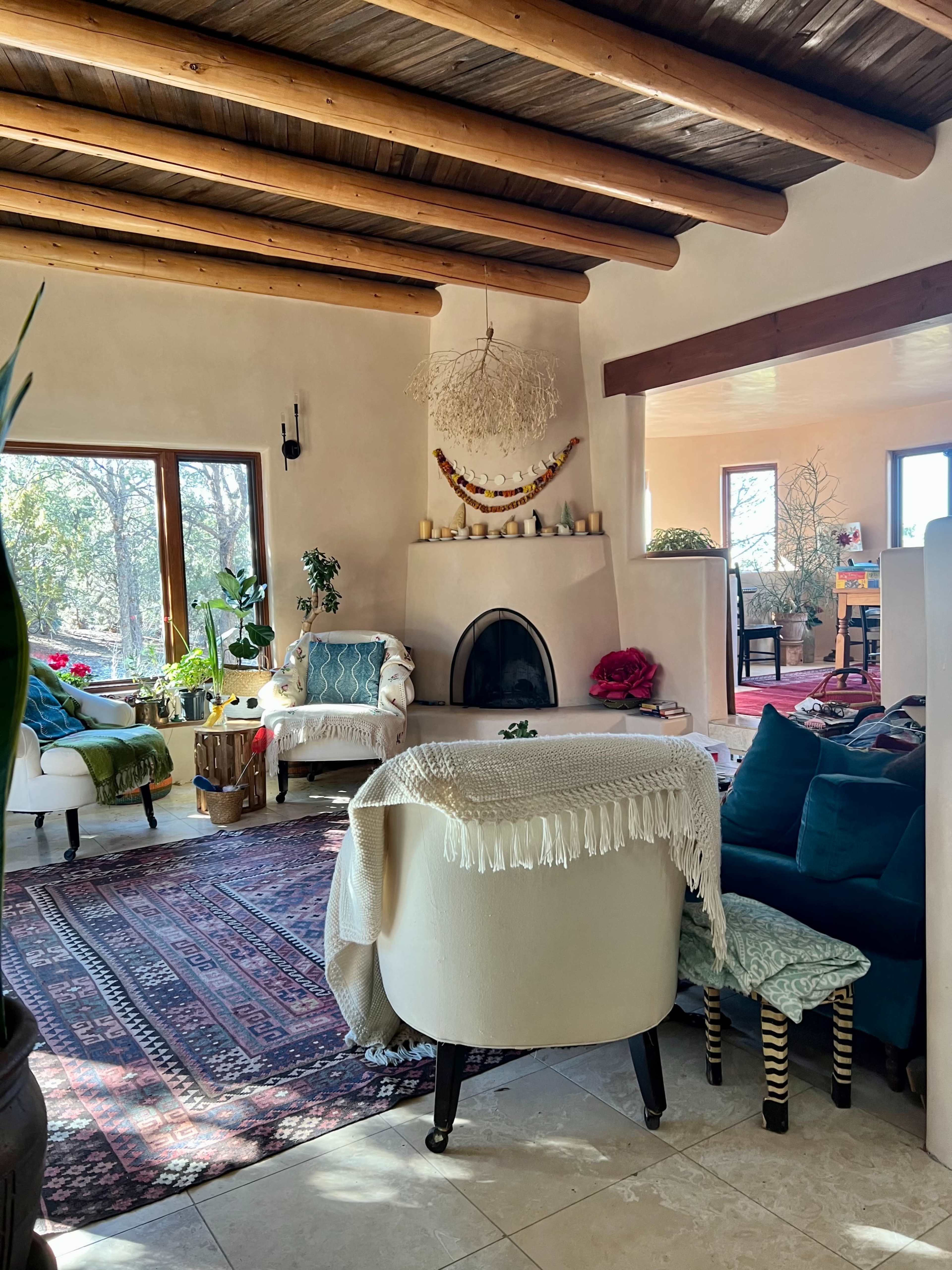 A cozy living room with a fireplace, wood-beamed ceiling, and a mix of seating arrangements, including a large white chair and various decorative elements.