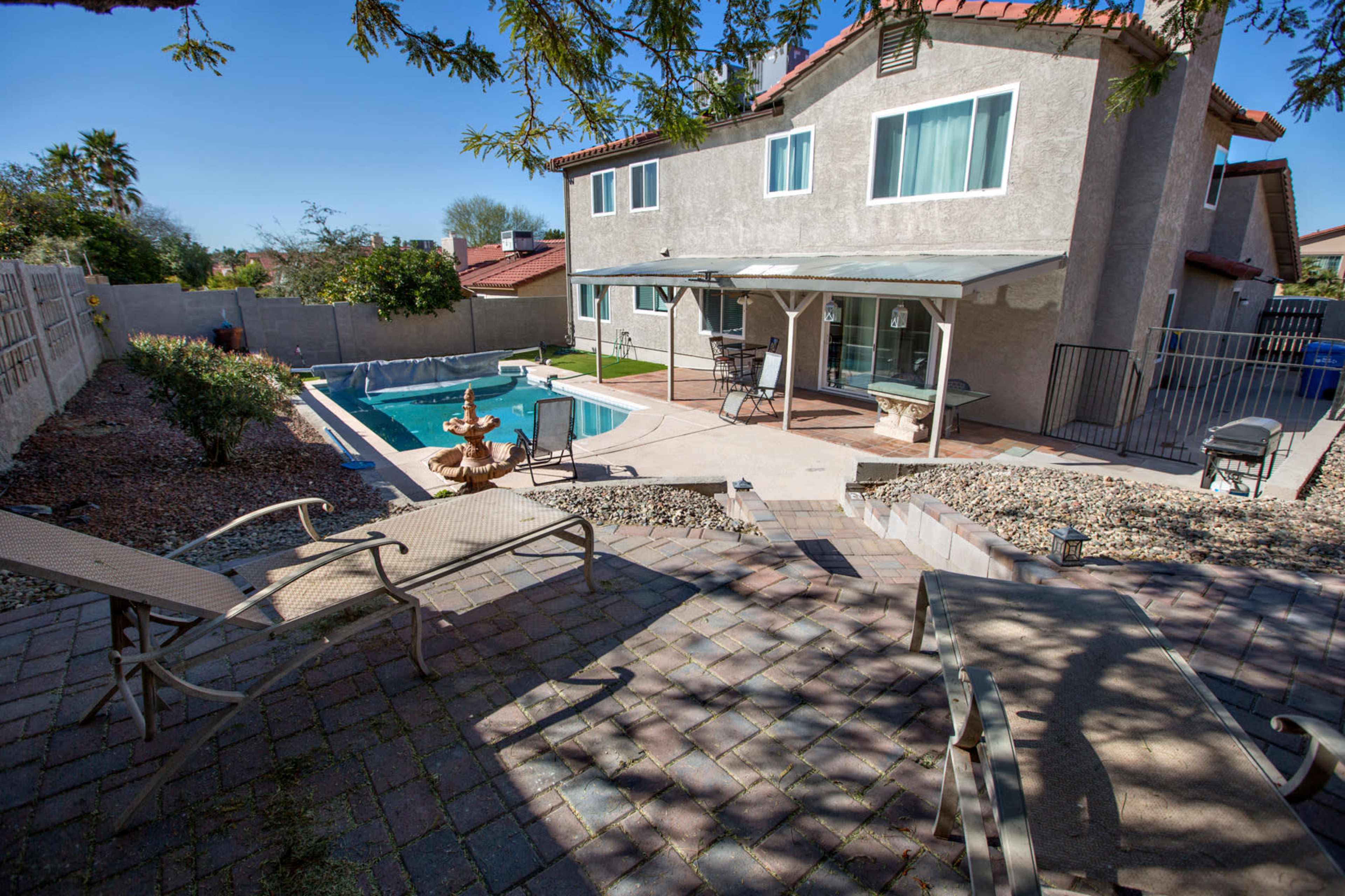 A backyard with a swimming pool, a fountain, and a two-story house bordered by a stone pathway and patio furniture.