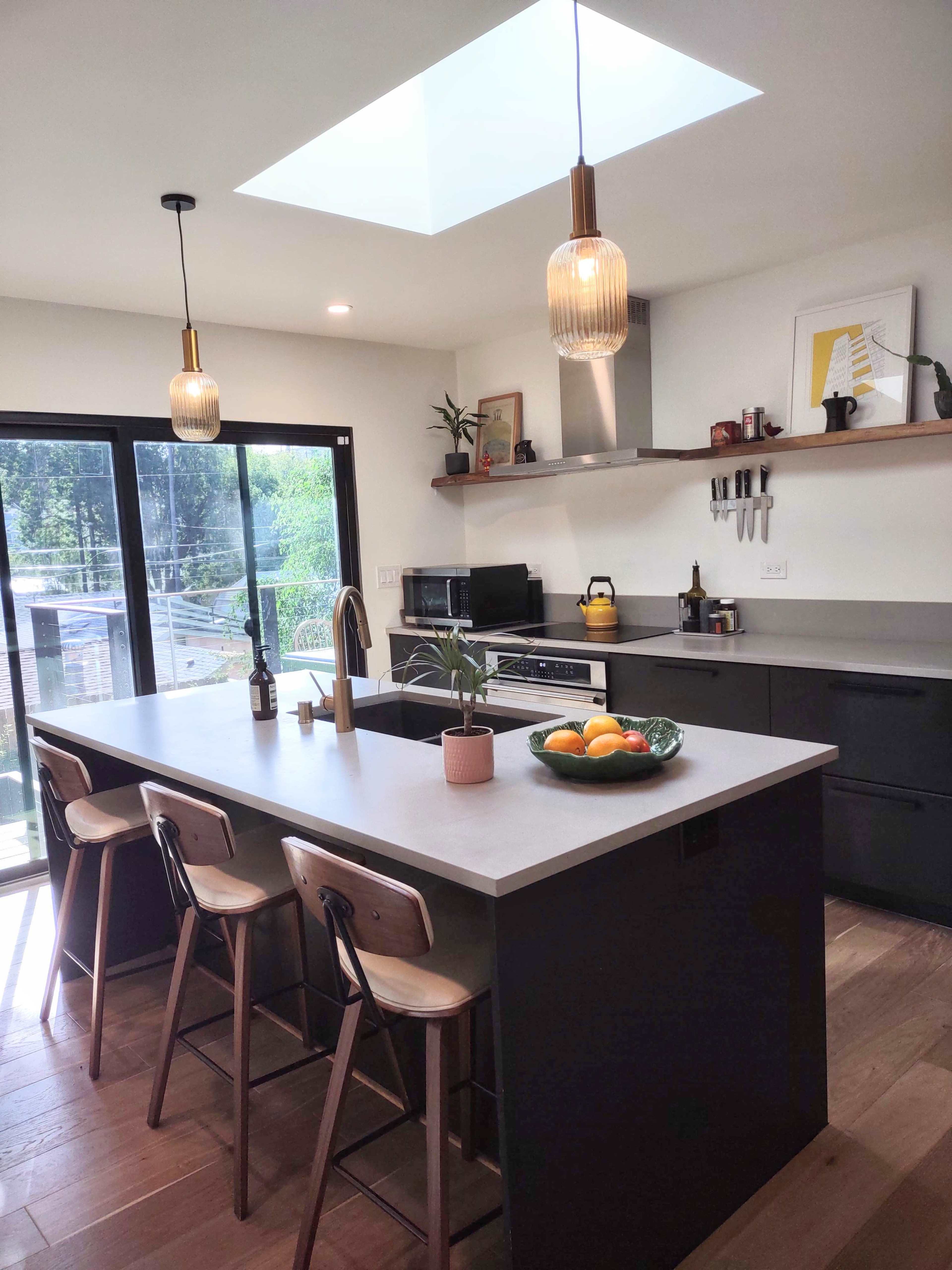 A modern kitchen features a central island with bar stools, pendant lights, a skylight, and a view of the outdoors.