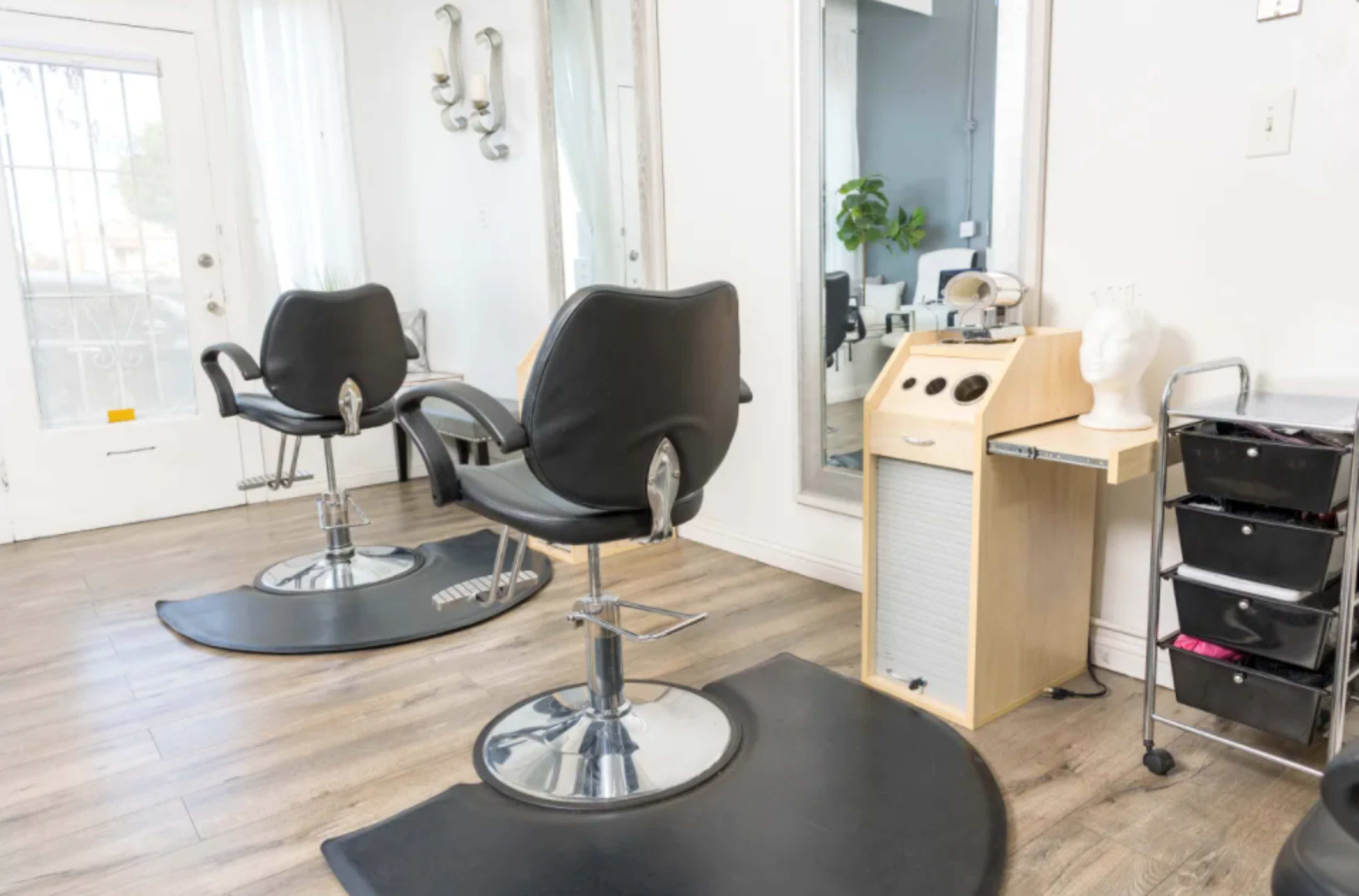A modern hair salon with two black styling chairs, a mirrored wall, and a small cart with storage drawers.