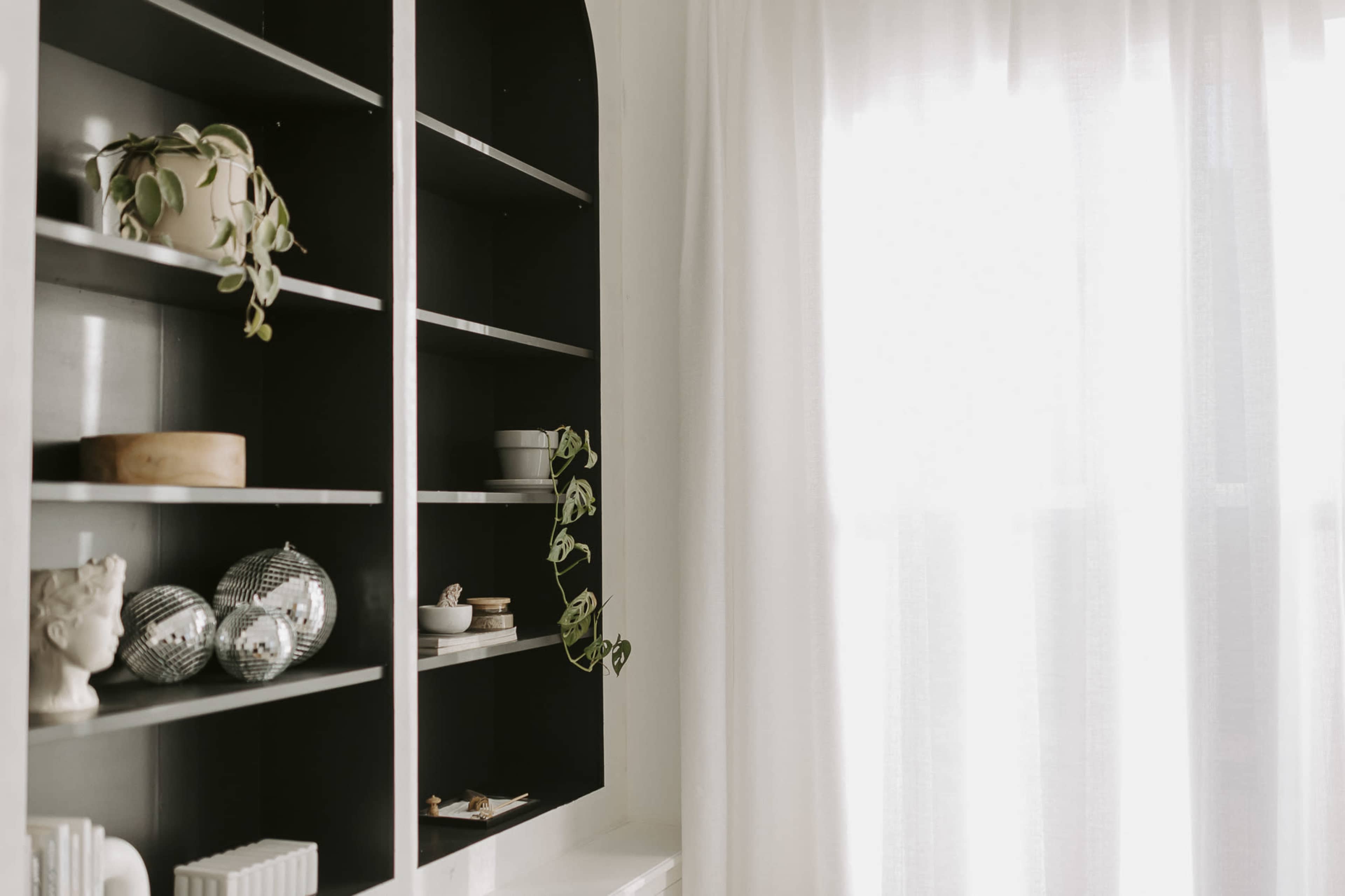 A black bookshelf with decorative items and plants is positioned beside a window with sheer white curtains.