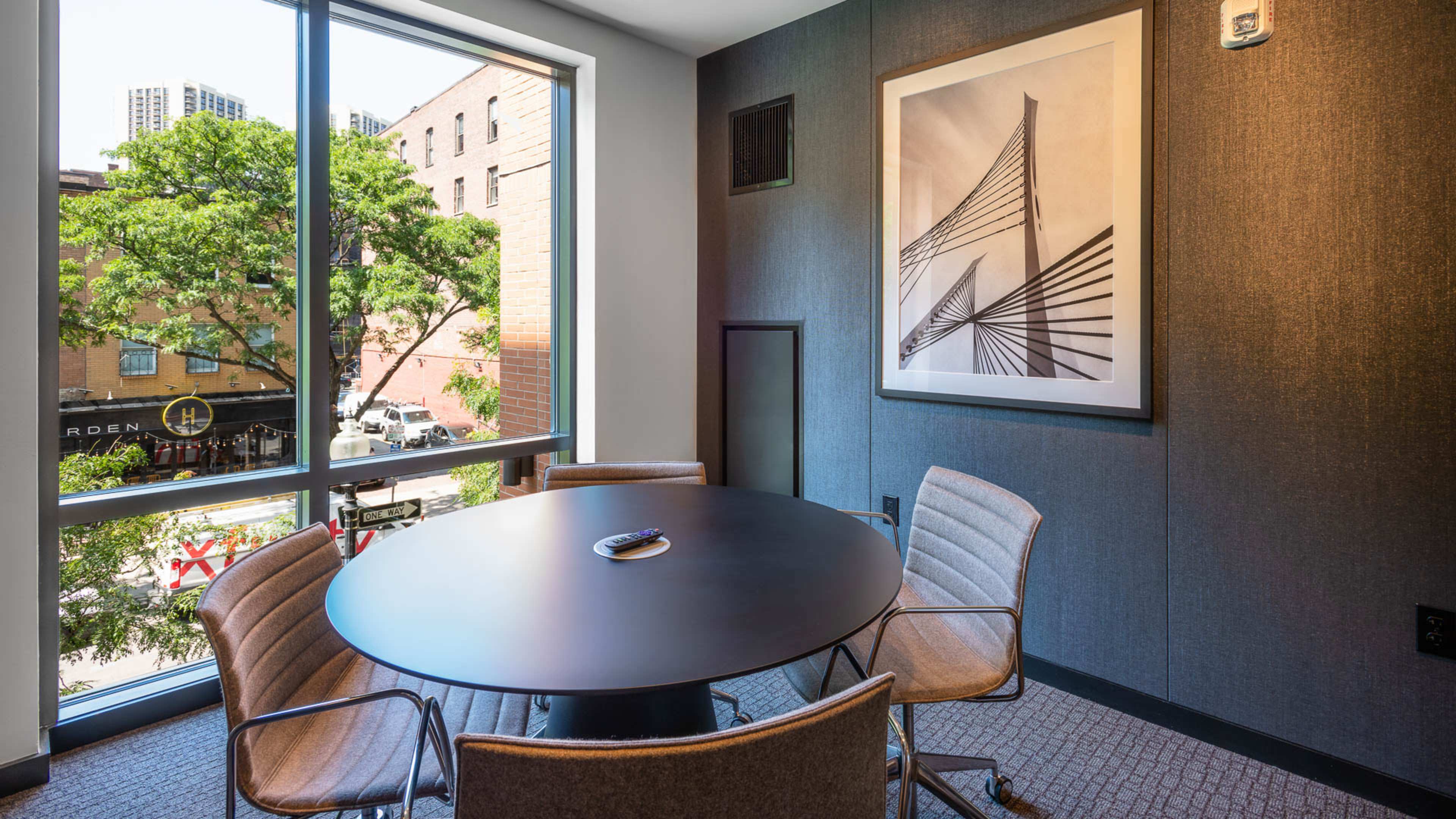A conference room features a round table surrounded by four chairs, with a large window displaying a view of the street and a framed artwork on the wall.