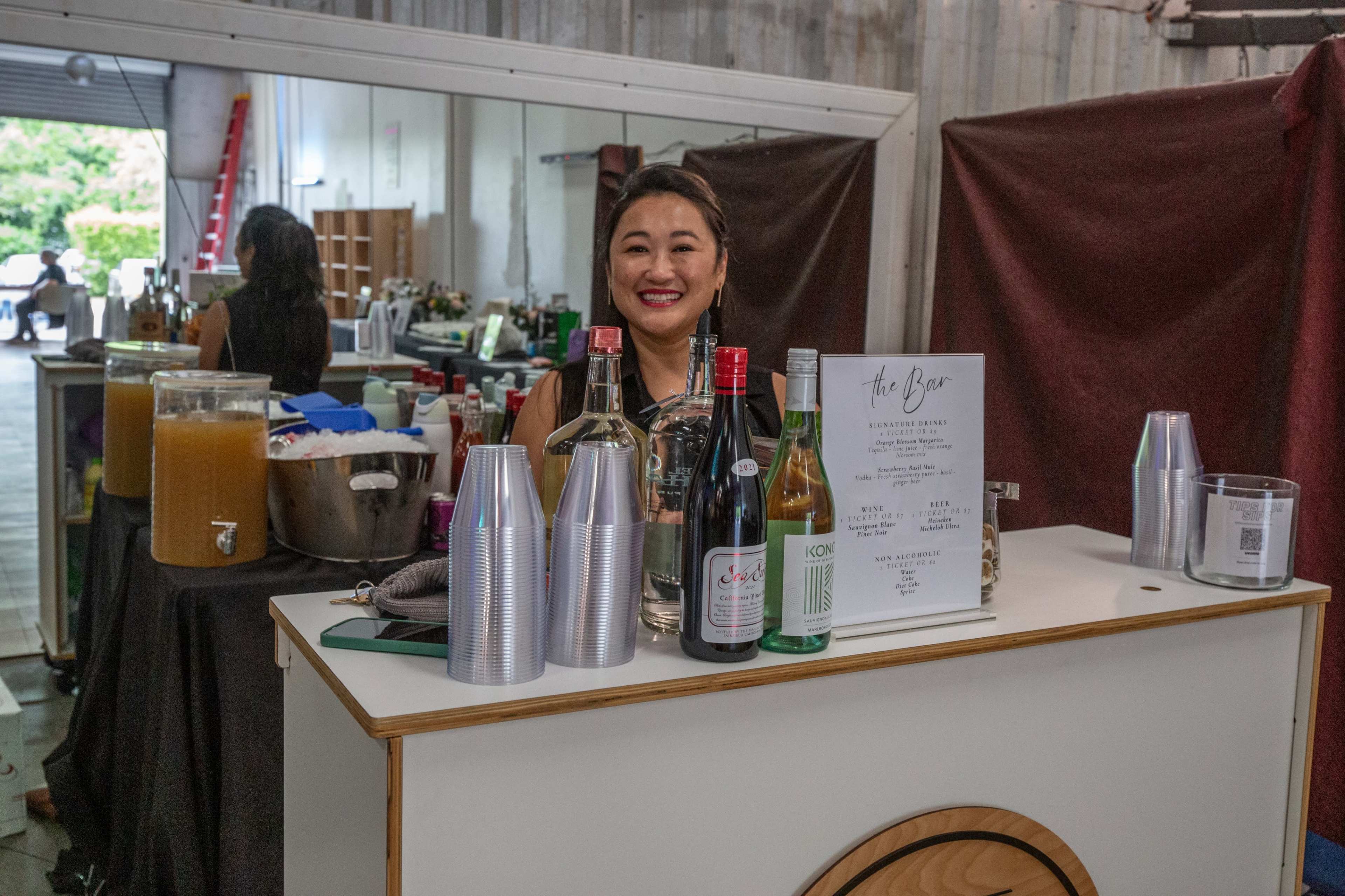 A woman stands behind a bar counter displaying various bottles of wine and spirits, with a drink menu visible nearby.