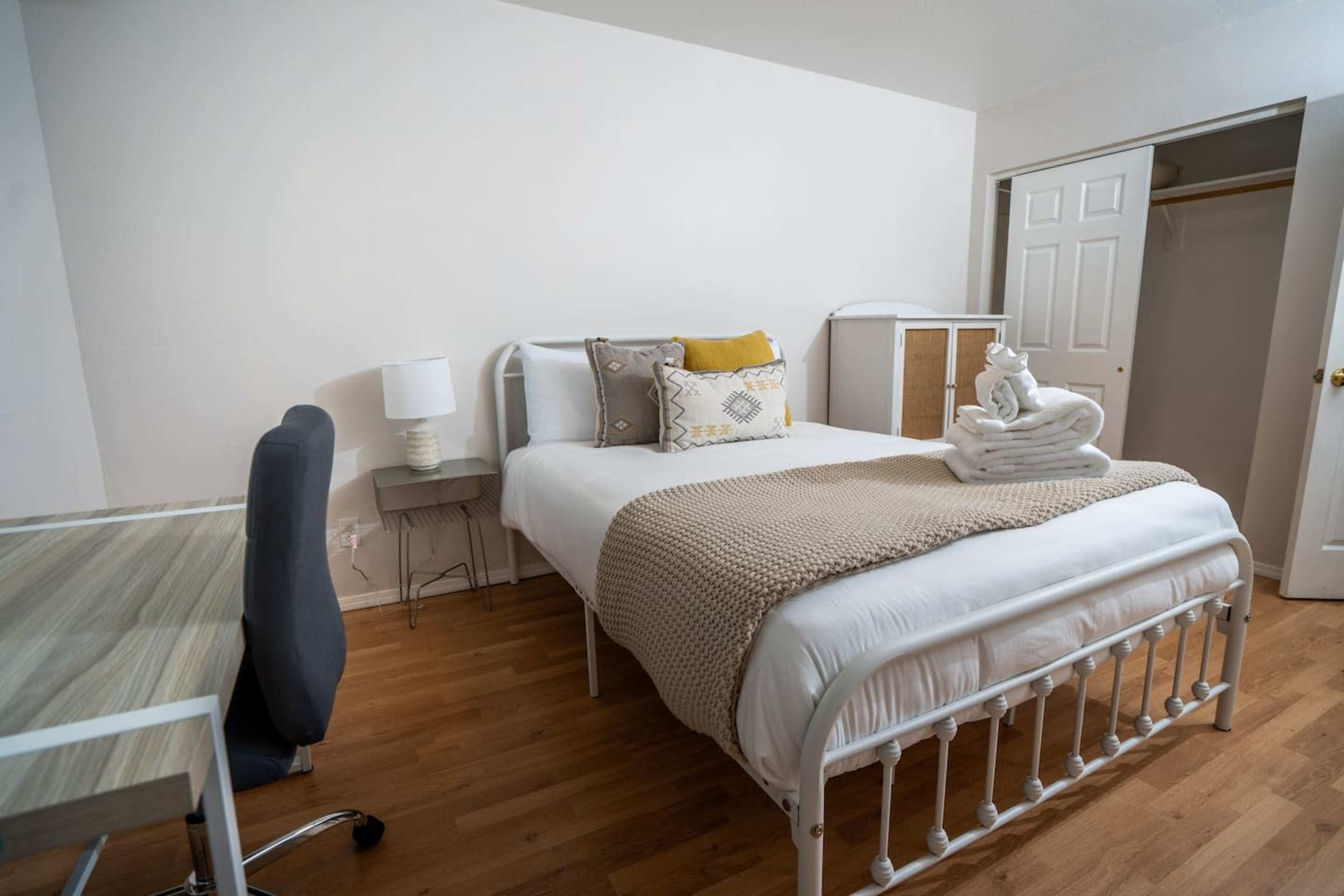 The image shows a neatly arranged bedroom featuring a bed with white linens and a decorative throw, a small desk with a chair, and a closet with sliding doors.
