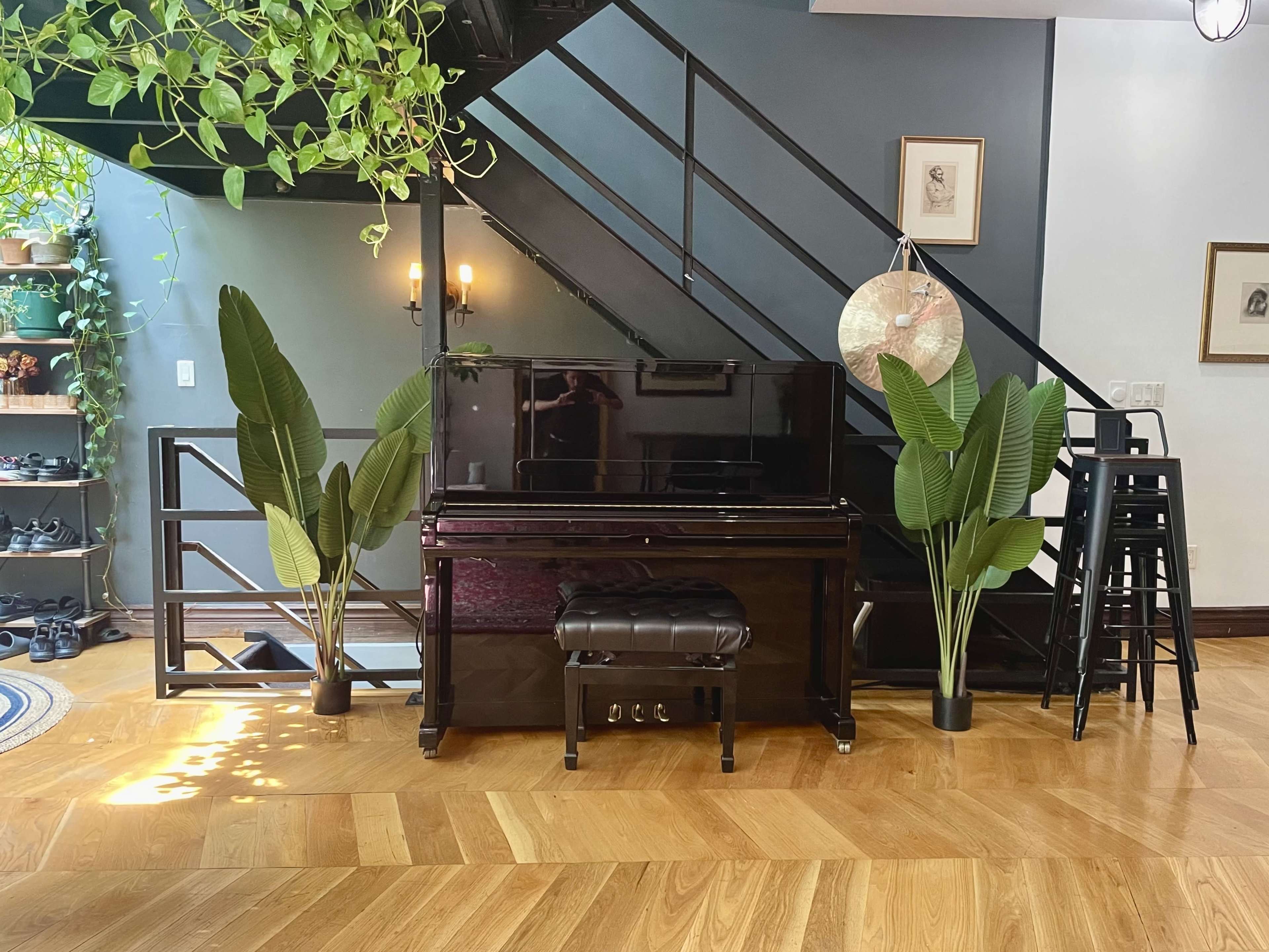 A black piano is positioned under a staircase, flanked by tall potted plants, with a small stool in front and a set of bar stools nearby.