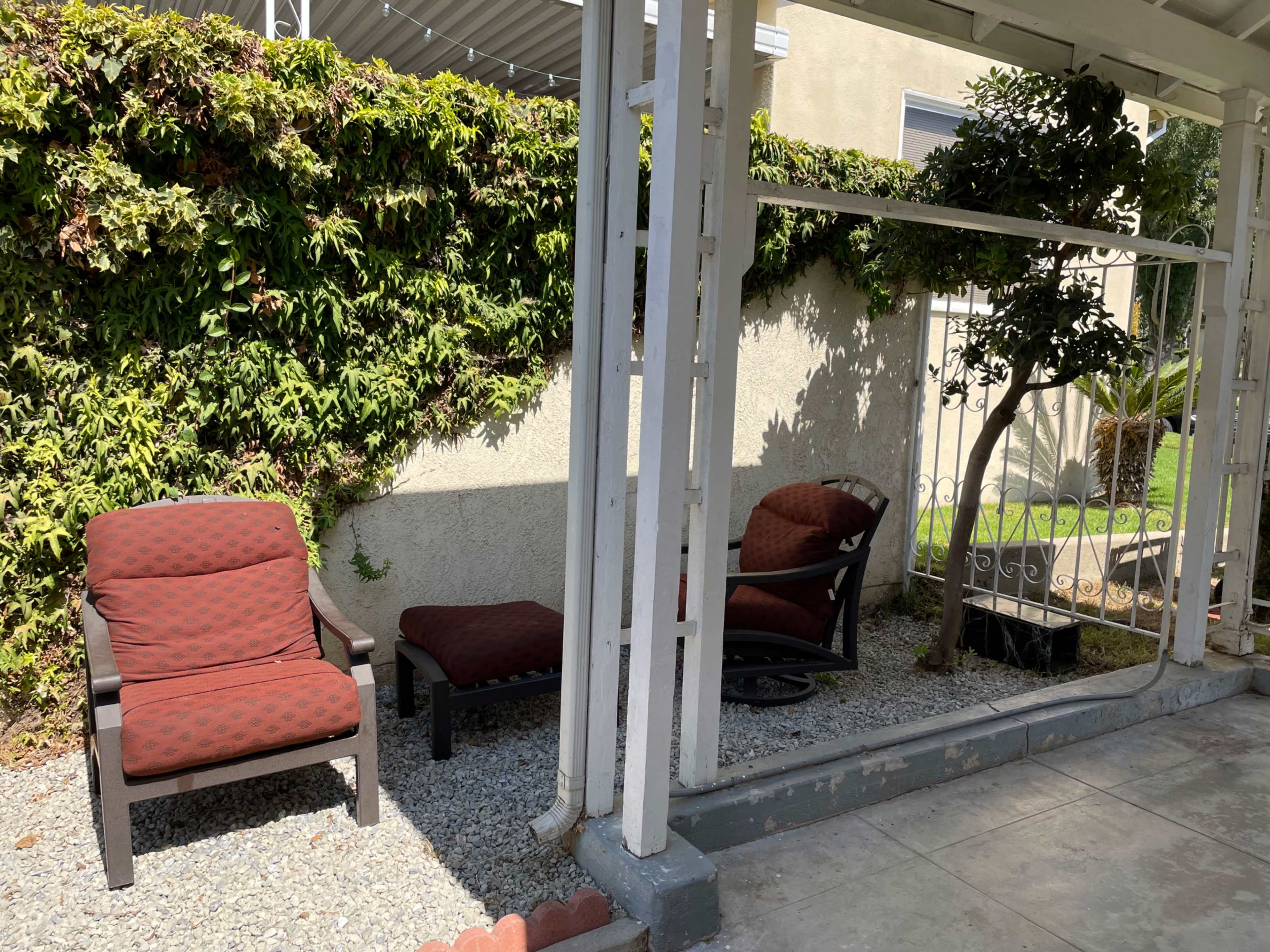 The image shows a small outdoor seating area with two chairs and a chaise lounge placed beside a green hedge and a tree.