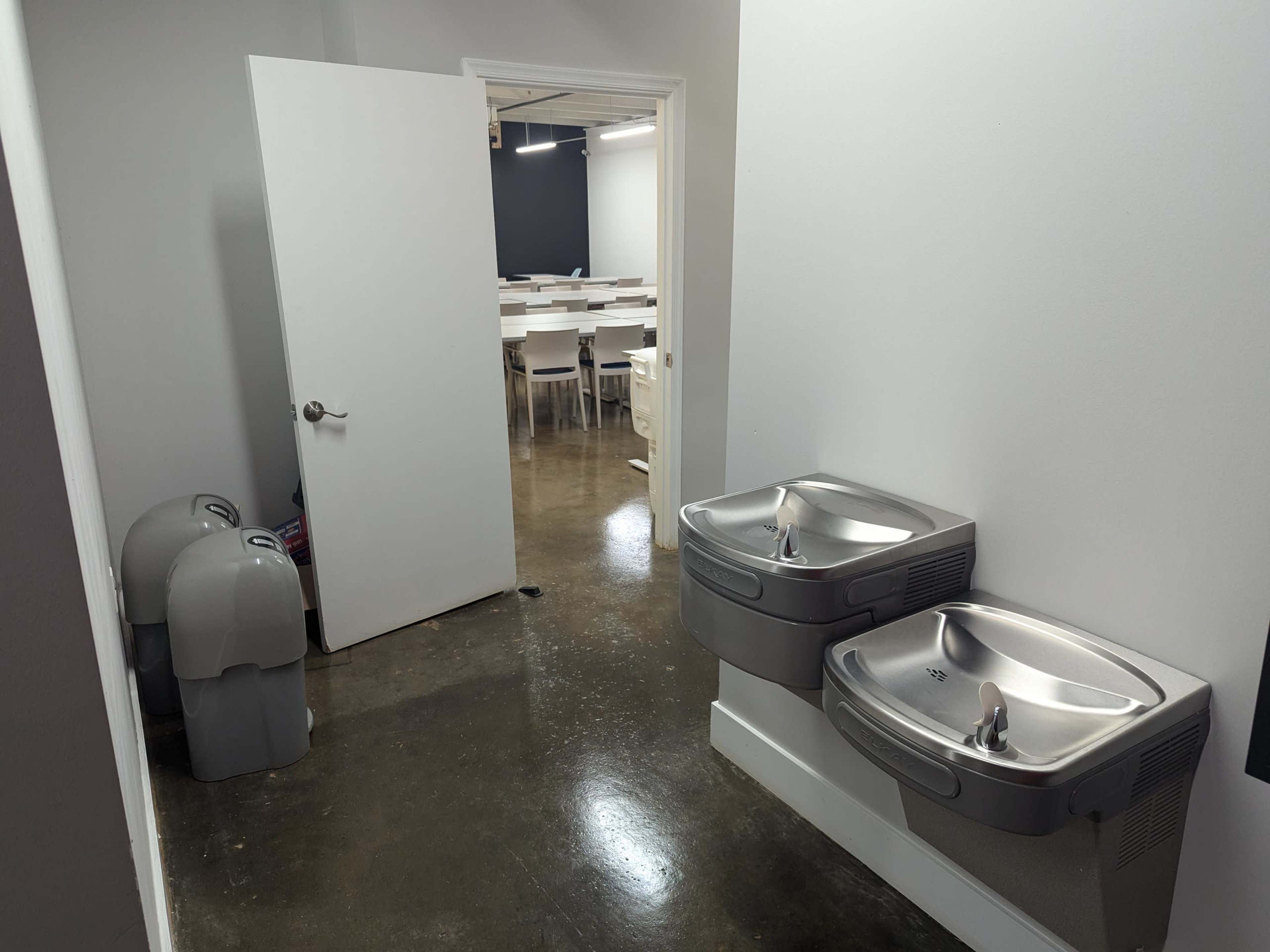 A hallway features two drinking fountains on one side and an open door leading to a classroom area in the background.