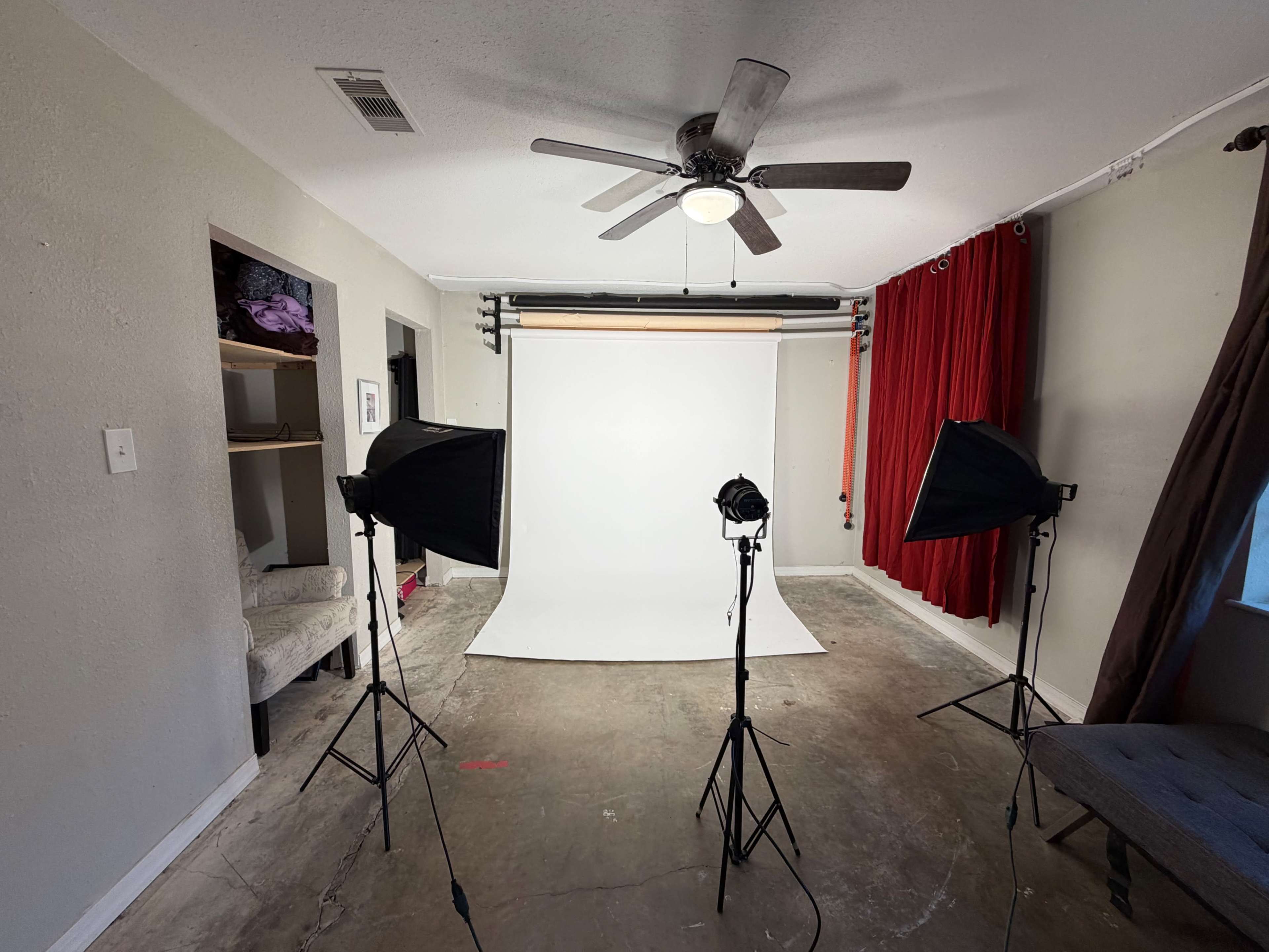 The image shows a photo studio setup with a white backdrop, two light sources, and red curtains in a room with a concrete floor.