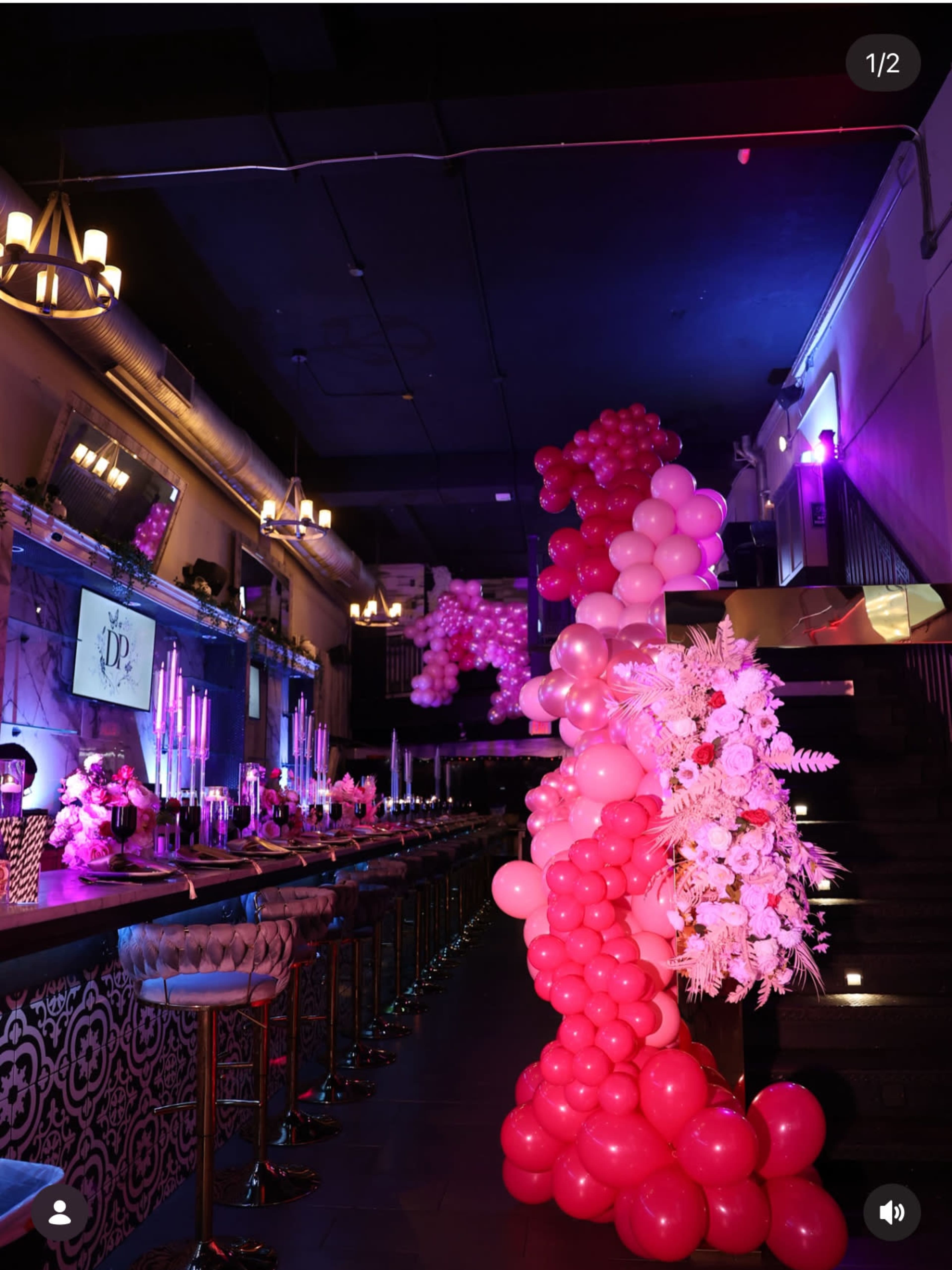 The image shows a dimly lit event space decorated with pink and white balloons, featuring a bar with candles and floral arrangements.