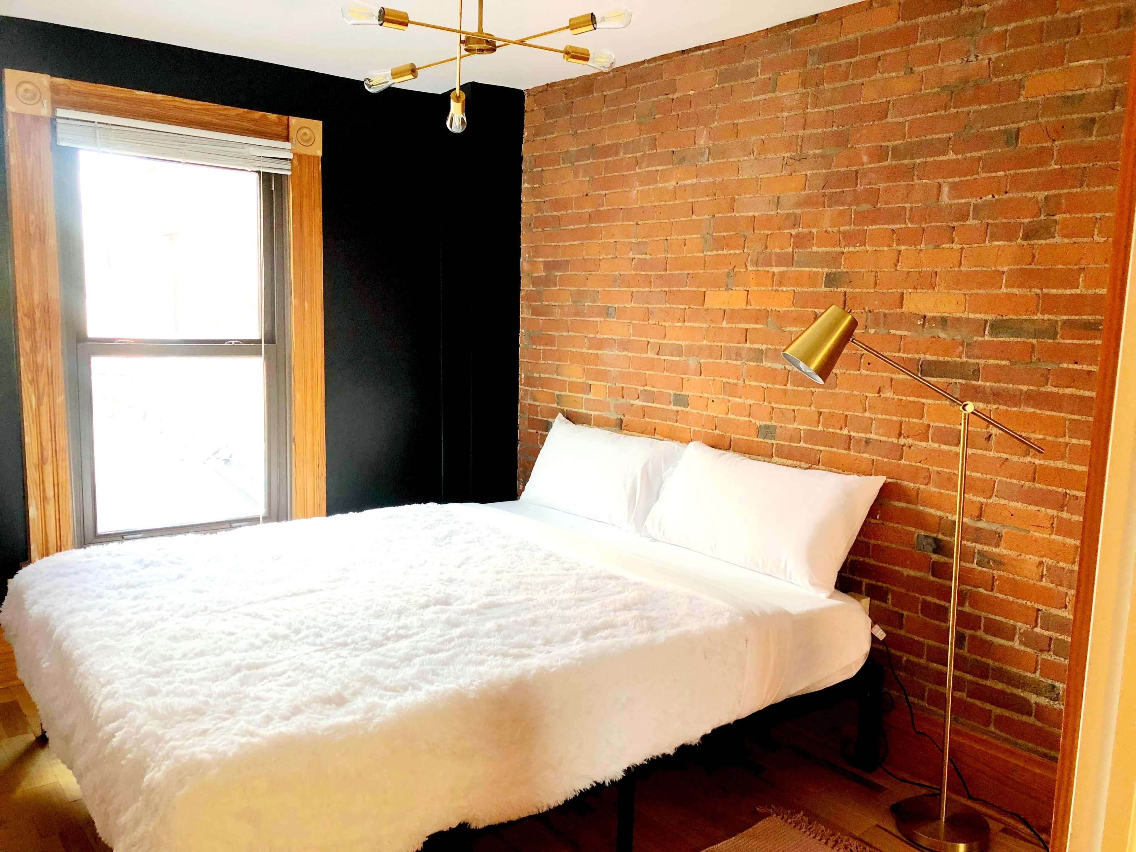 A modern bedroom featuring a bed with a white bedspread, a gold floor lamp, and exposed brick walls.