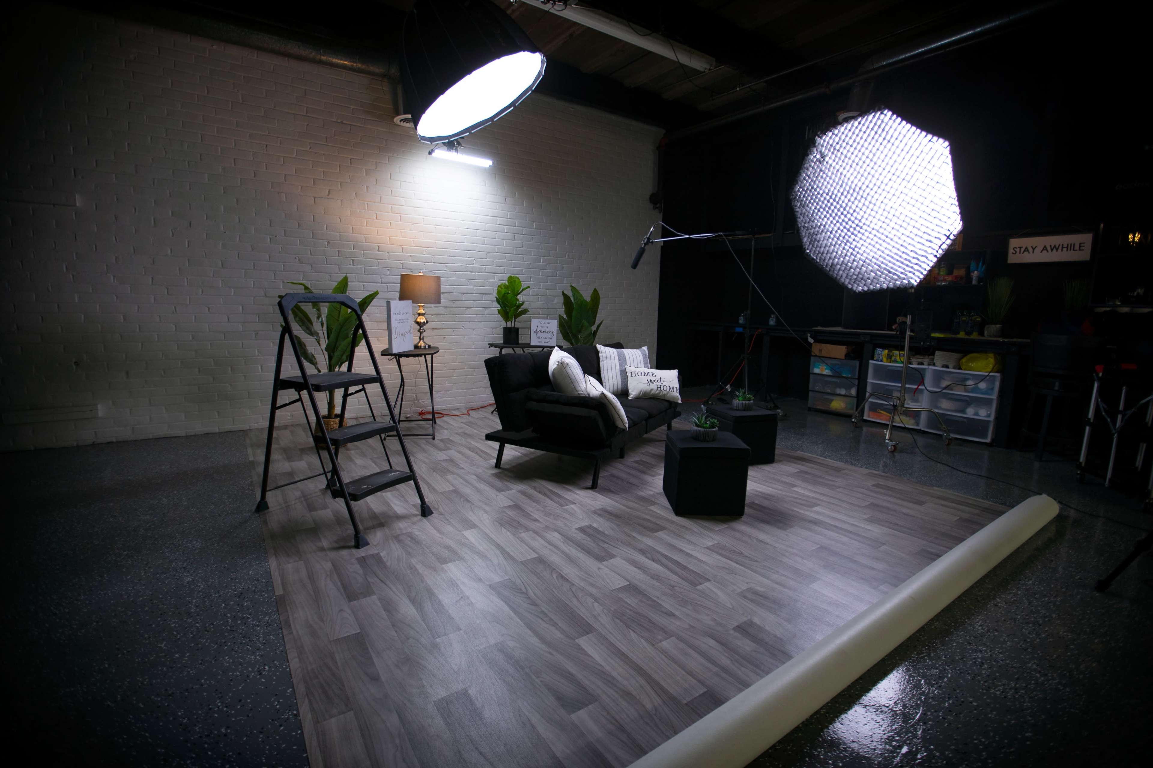 A staged indoor setting with two black chairs, a small table, plants, and lighting equipment on a wooden floor.