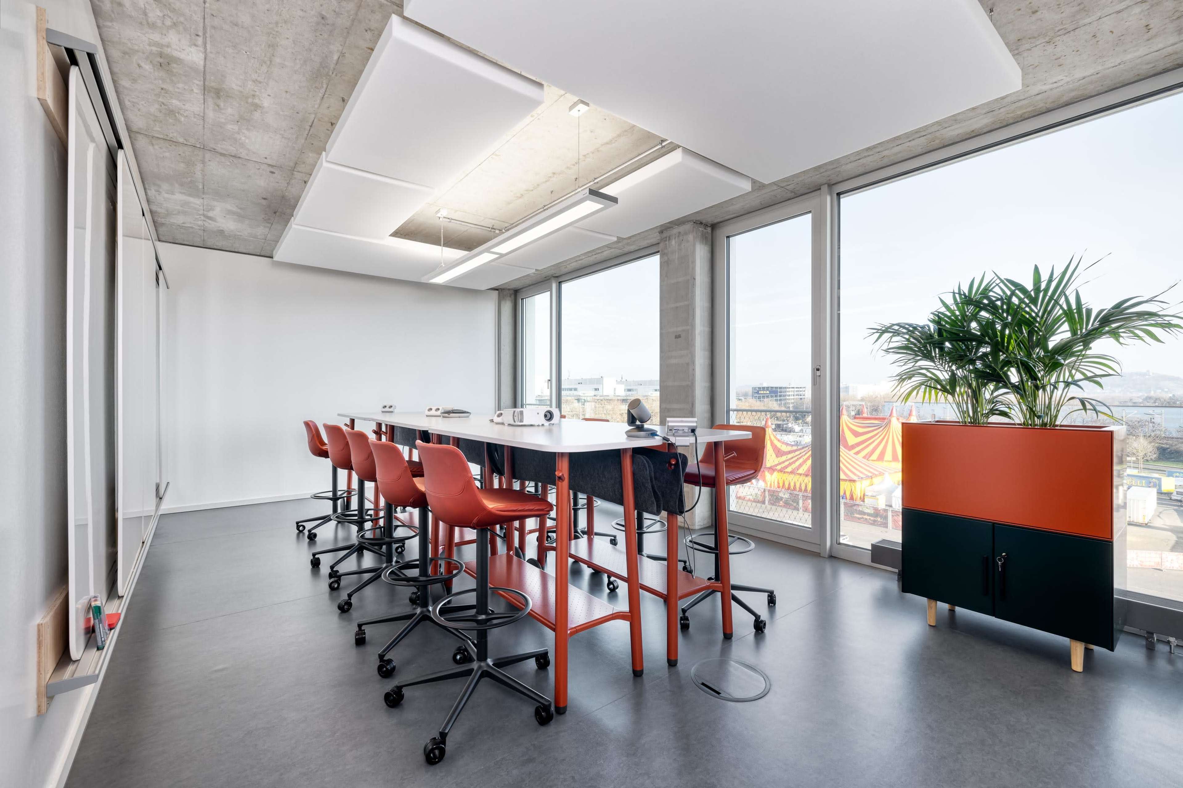 A modern office space featuring a large table with high stools, floor-to-ceiling windows, and a plant beside a colorful storage cabinet.