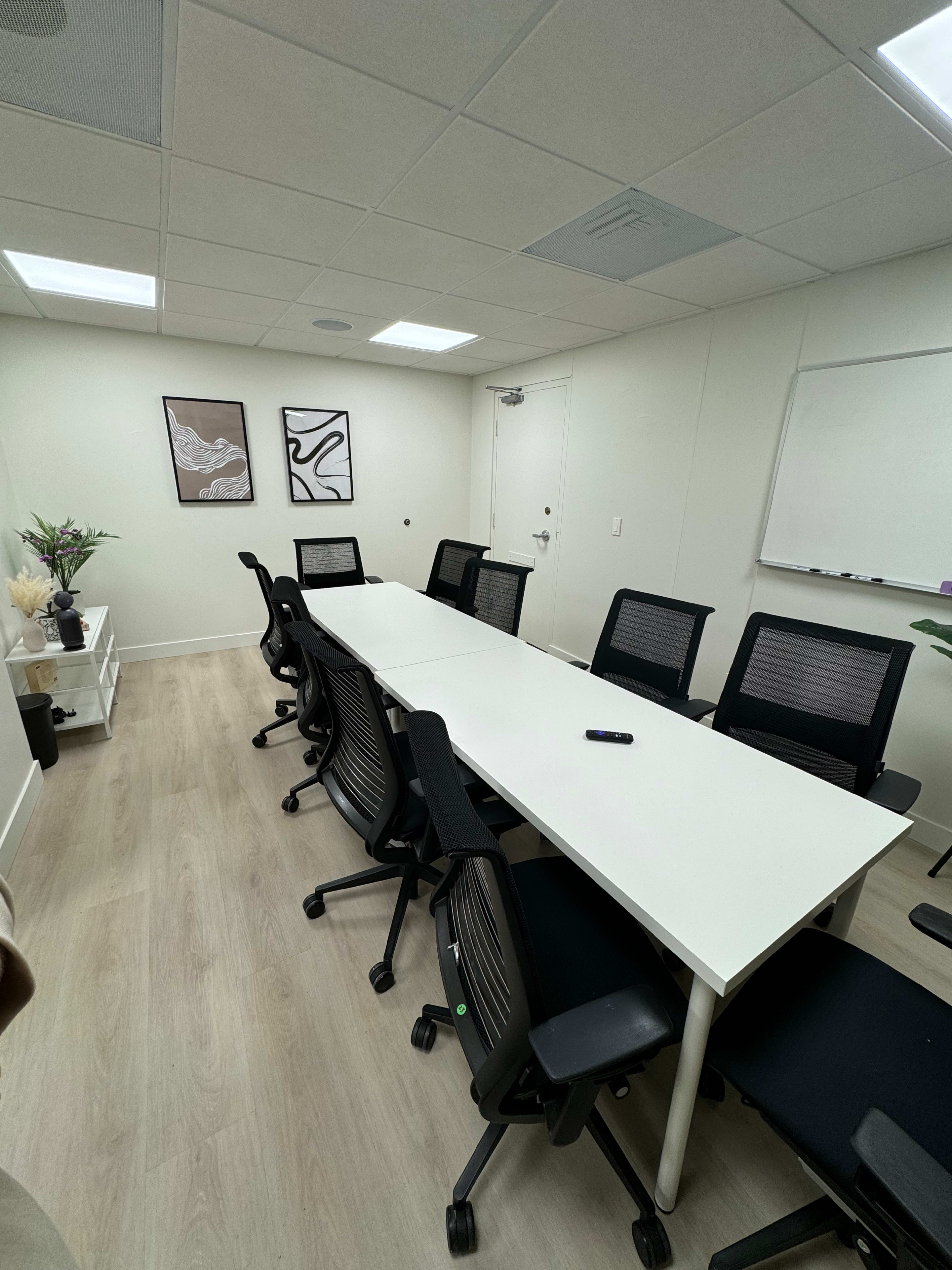 A modern conference room with a long white table surrounded by black office chairs, a whiteboard on the wall, and two framed artworks.