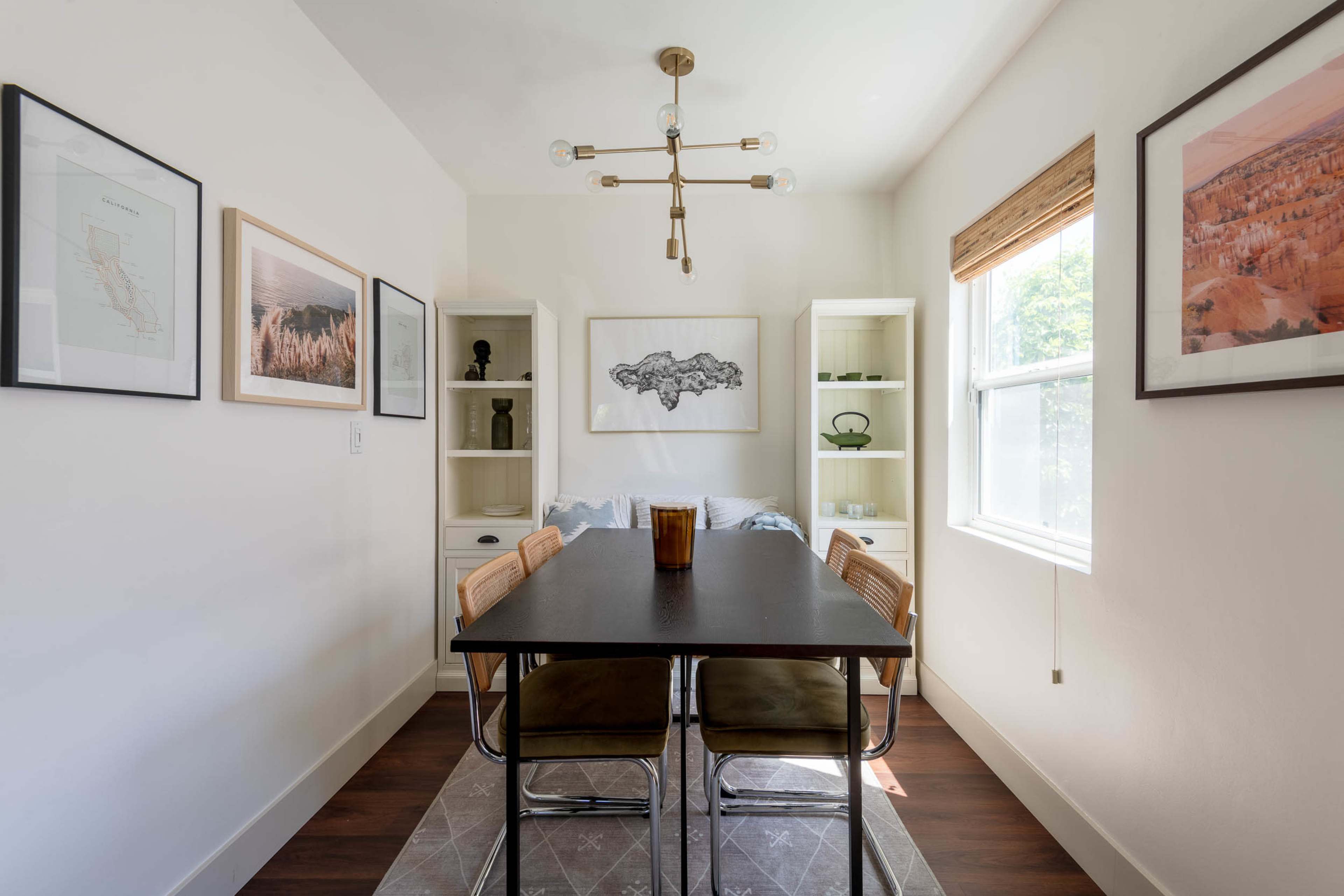 The image shows a modern dining area with a wooden table surrounded by chairs, light-colored walls, and framed artwork hanging on the walls.