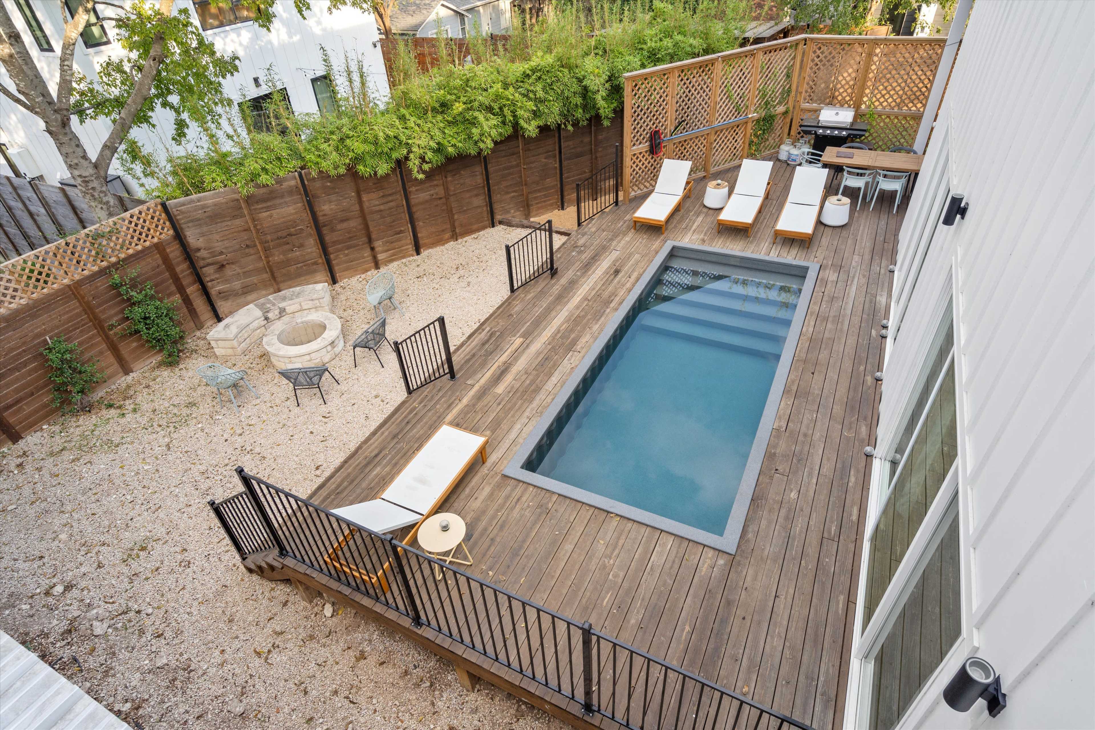 The image shows an aerial view of a wooden deck with a rectangular swimming pool, surrounded by lounge chairs, a fire pit, and outdoor seating in a backyard.