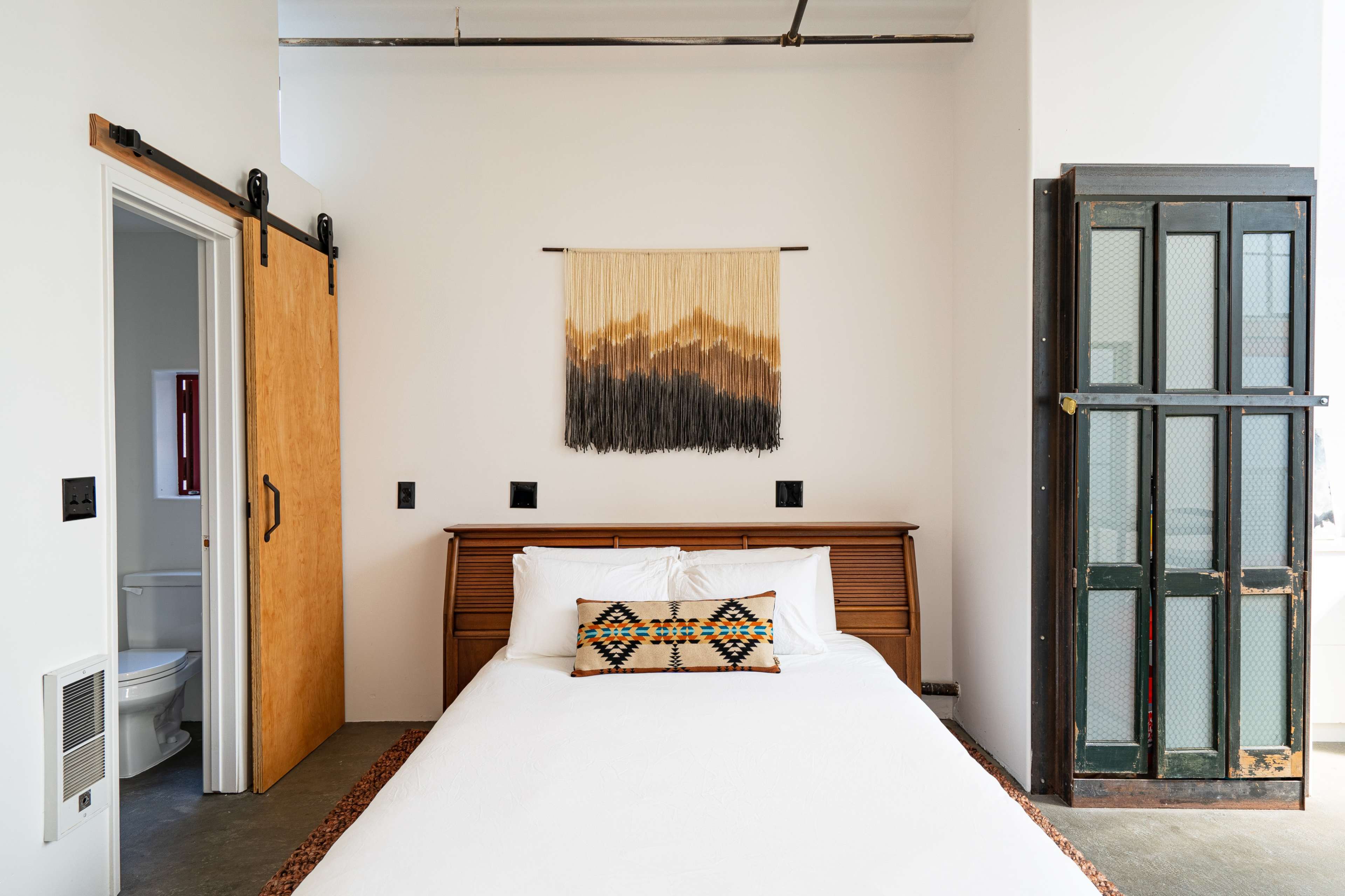 A modern bedroom featuring a king-sized bed with a patterned pillow, a wall hanging above, a wooden sliding door, and a vintage-style glass door on one side.