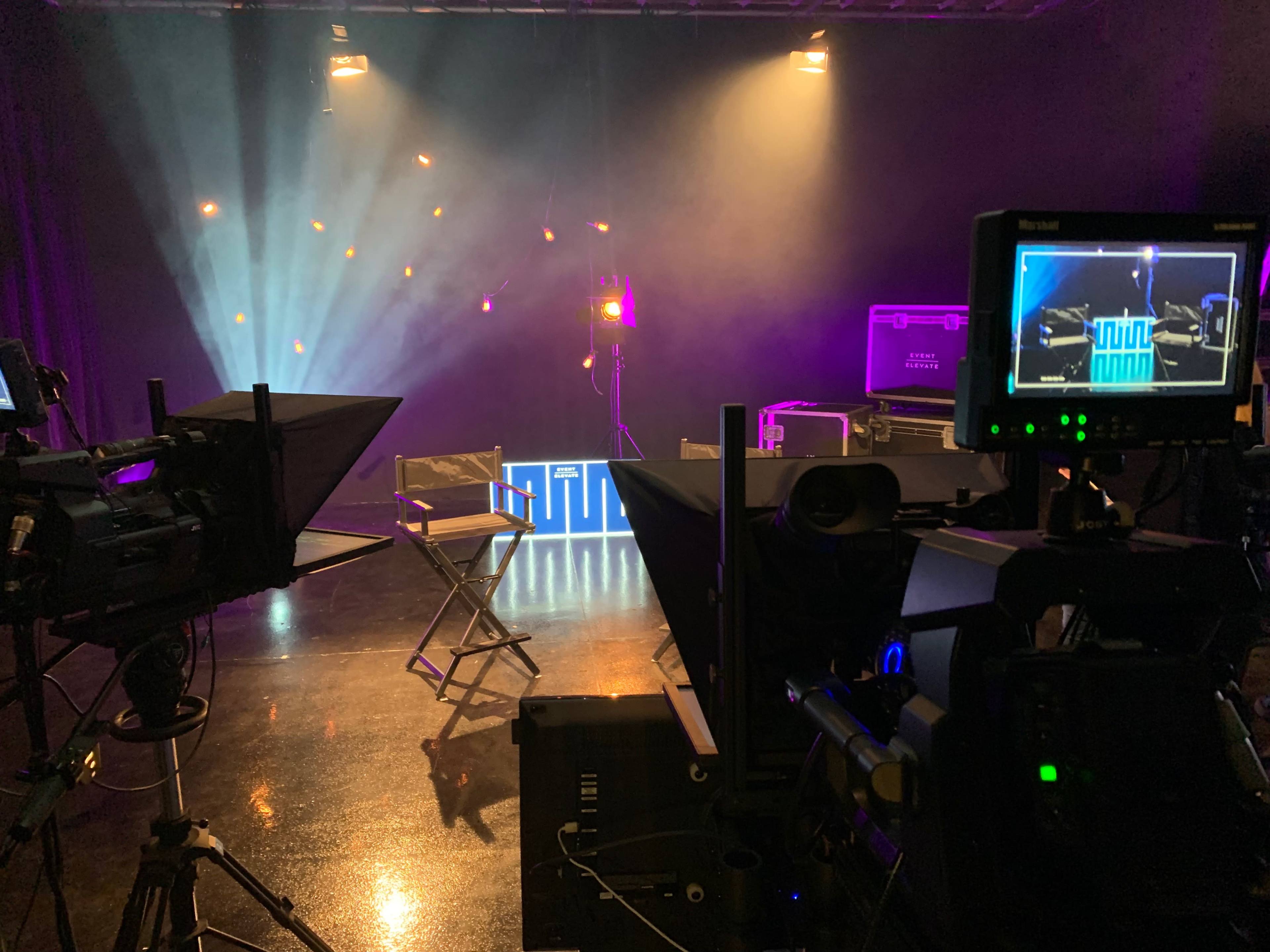 A dimly lit studio setup with cameras and equipment directed towards a stage featuring colorful lights and a chair.