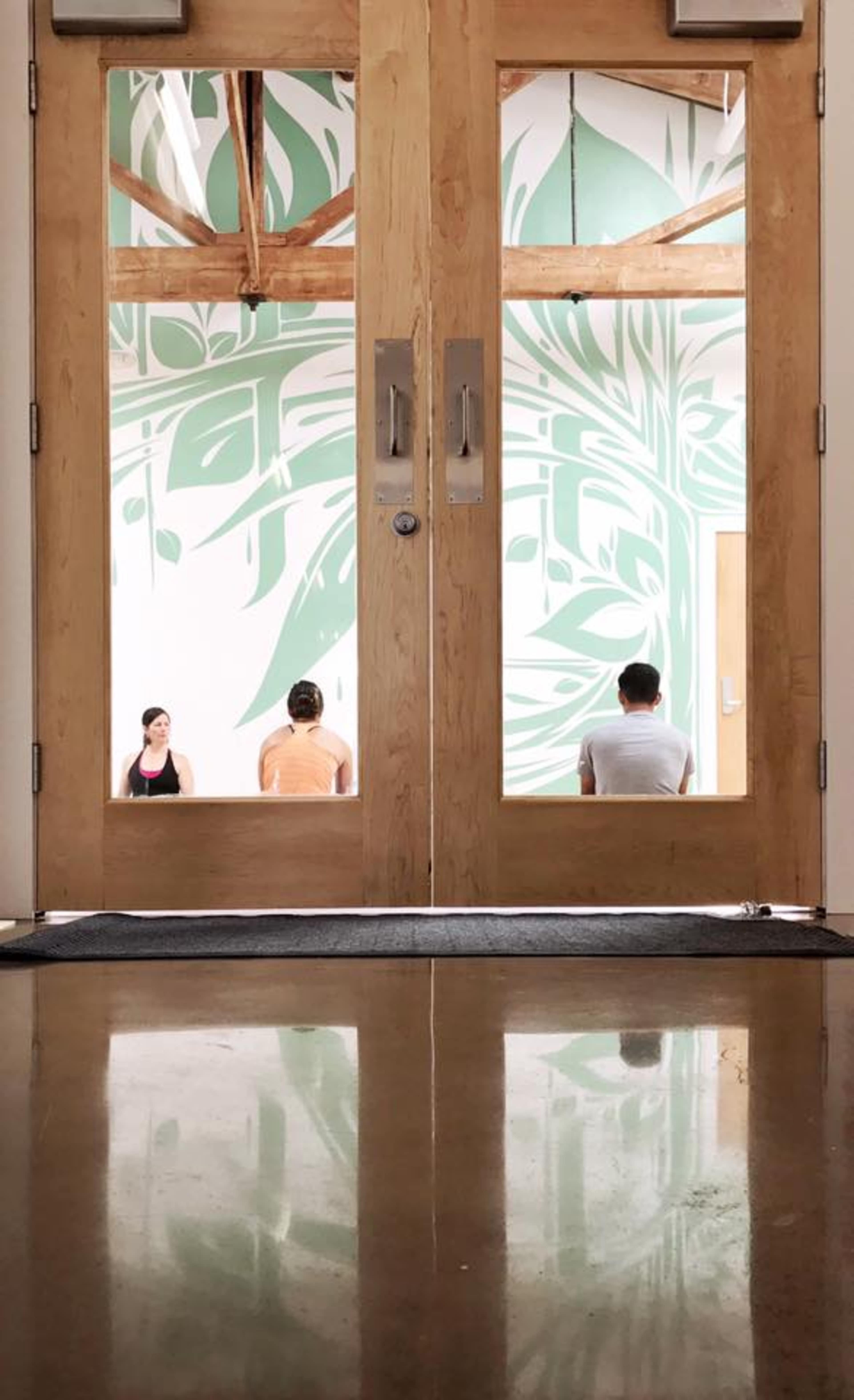 Two individuals sit in a yoga studio with a large green leaf mural on the wall, viewed through wooden double doors.