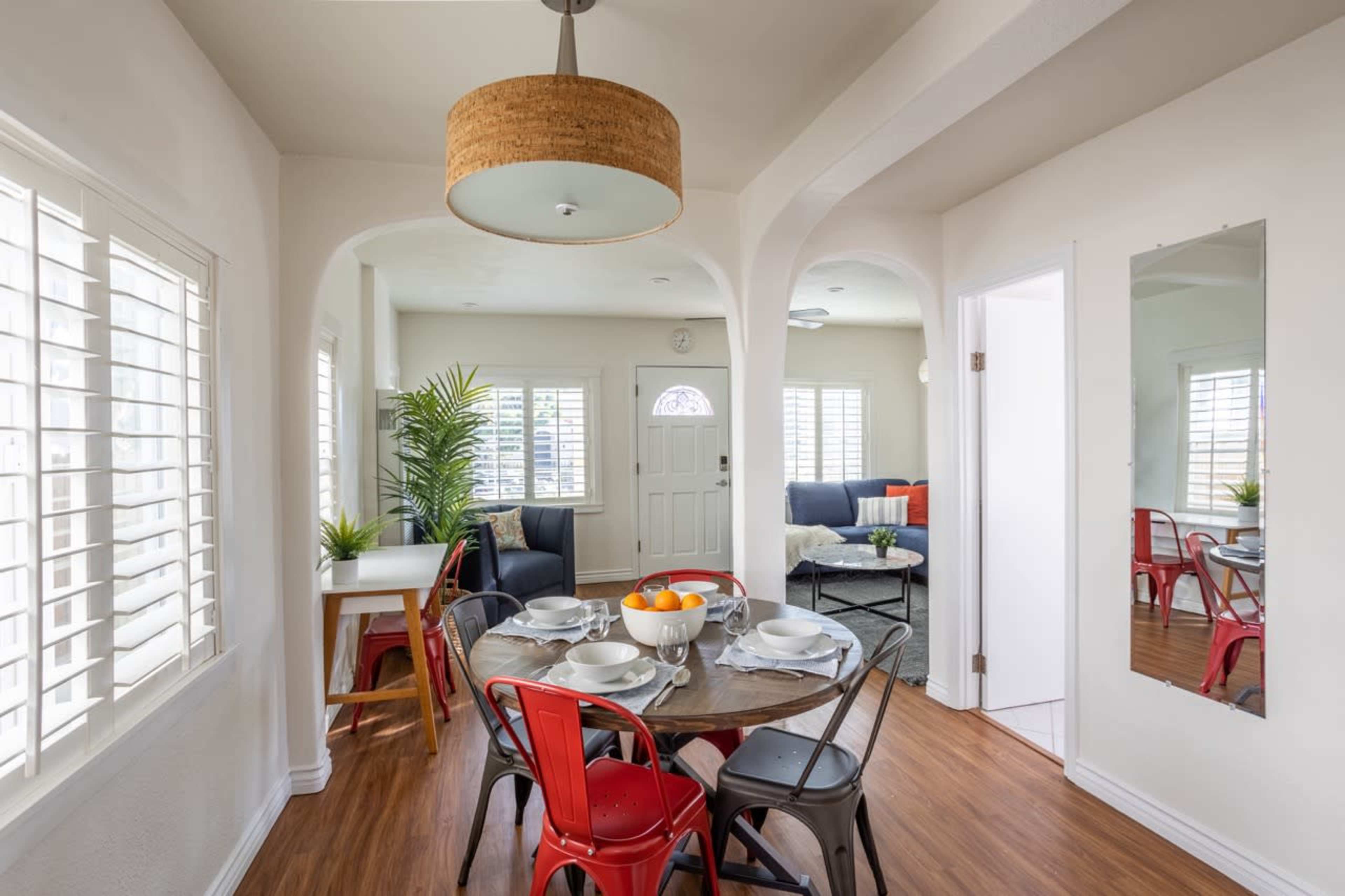 A dining area features a round table set with dishes and surrounded by red and black chairs, adjacent to a living space with a large couch and green plants under natural light from windows.