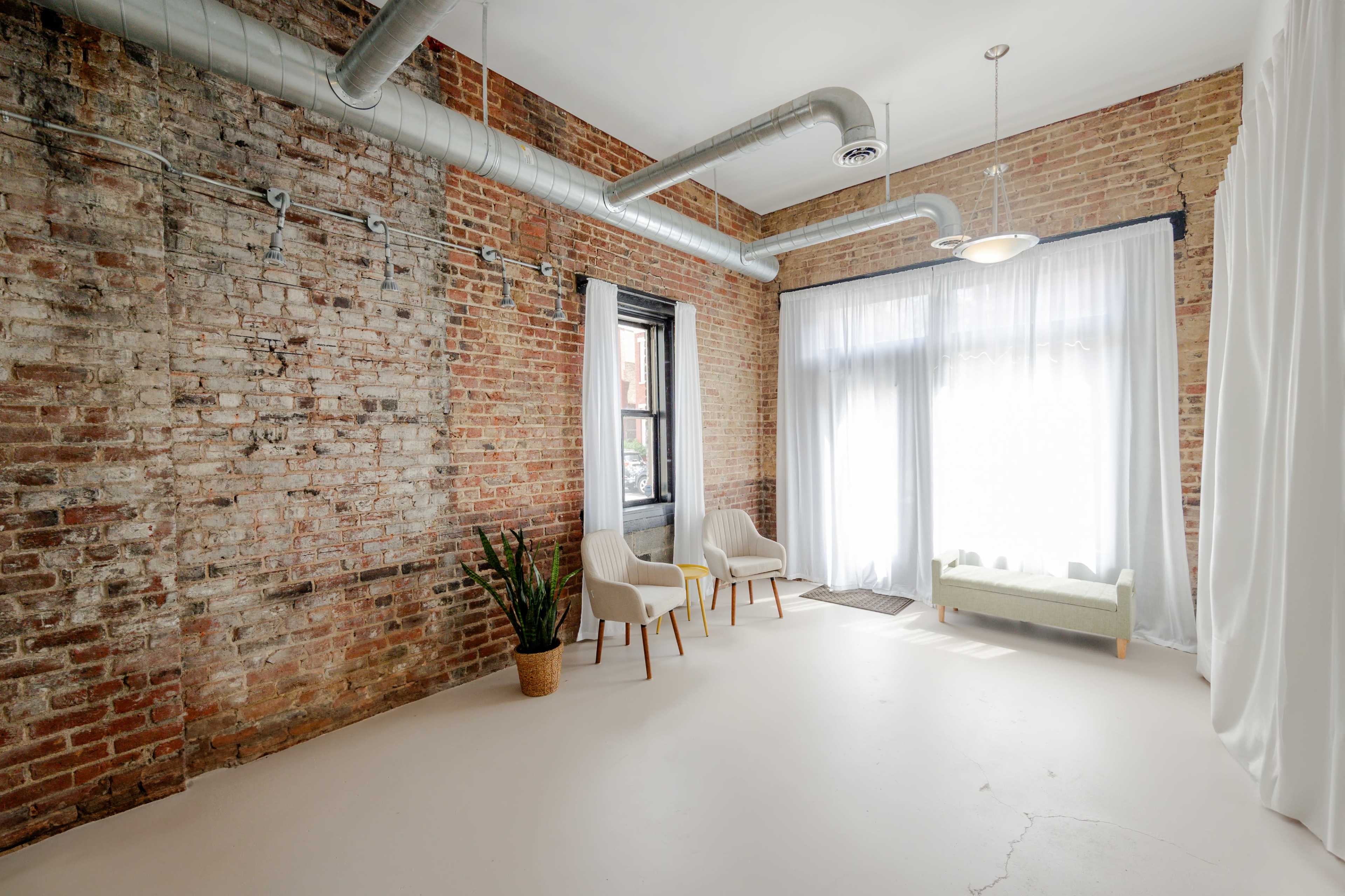 A spacious room with exposed brick walls, two chairs, a small couch, and large windows covered by sheer curtains.