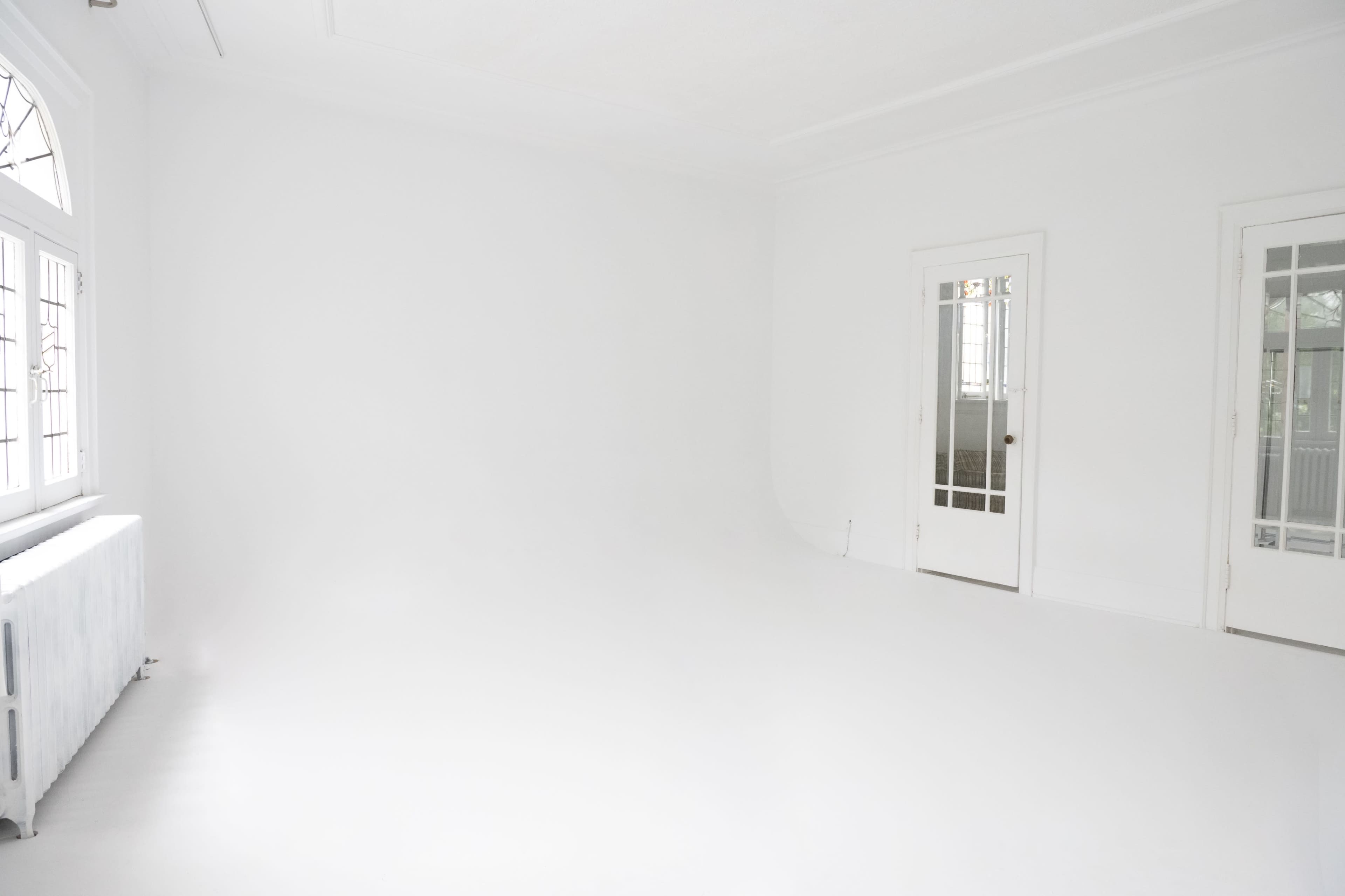 A spacious, empty room with white walls and a white floor, featuring large windows and a door leading to another room.