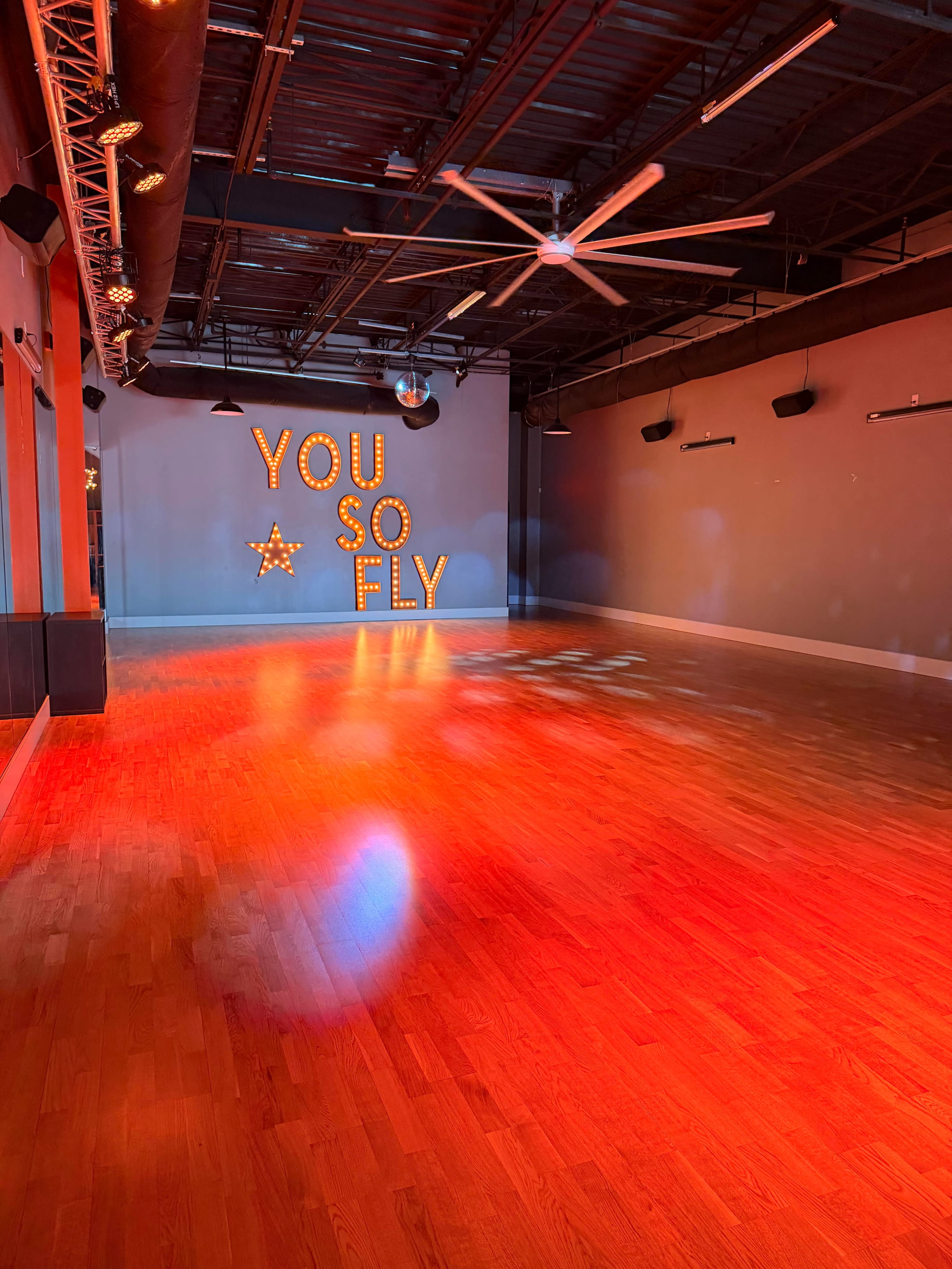 An empty dance studio features wooden floors and a large illuminated sign that reads "YOU SO FLY."