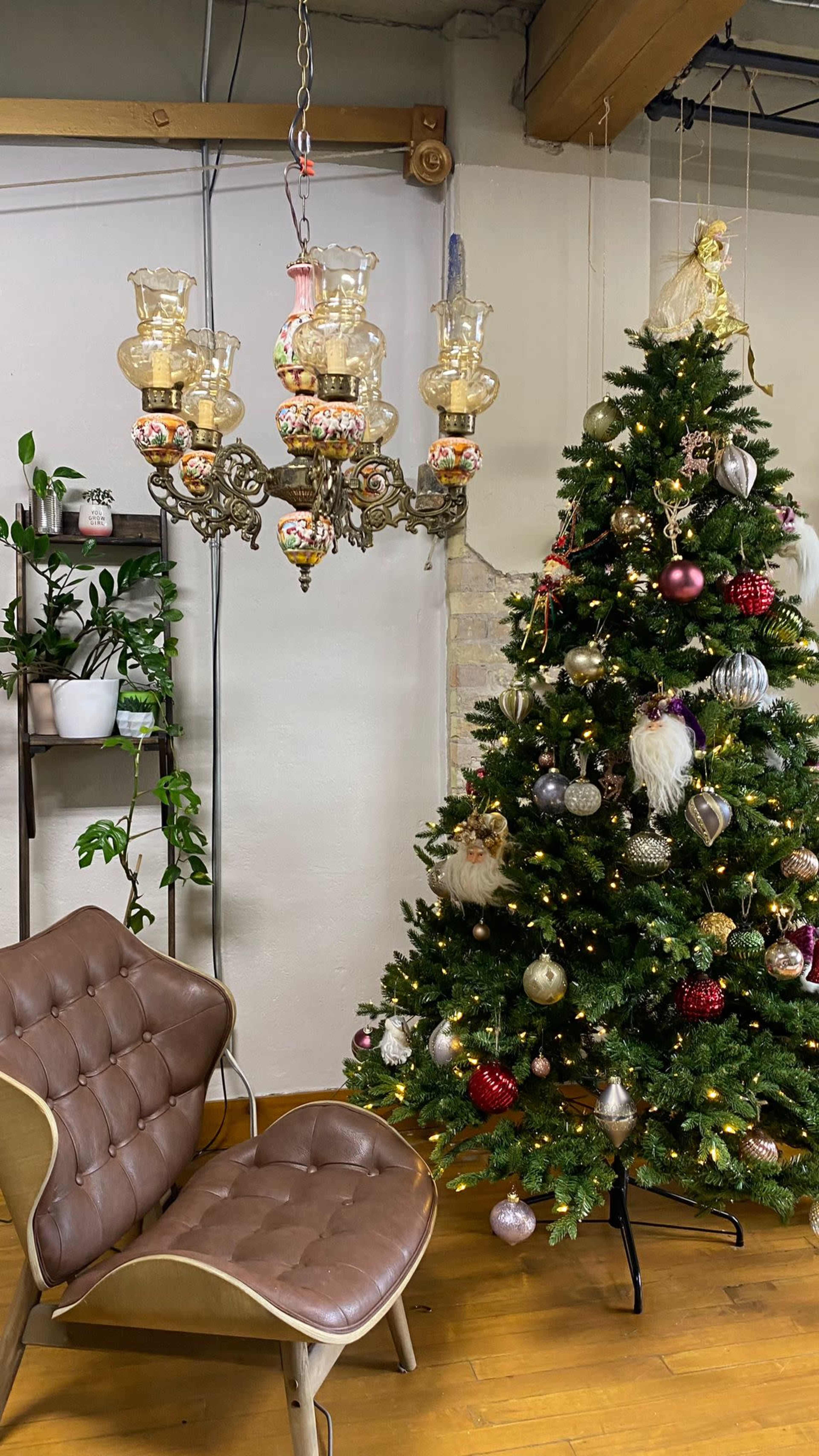 A decorative chandelier hangs above a Christmas tree adorned with ornaments, next to a brown leather chair in a cozy indoor space.