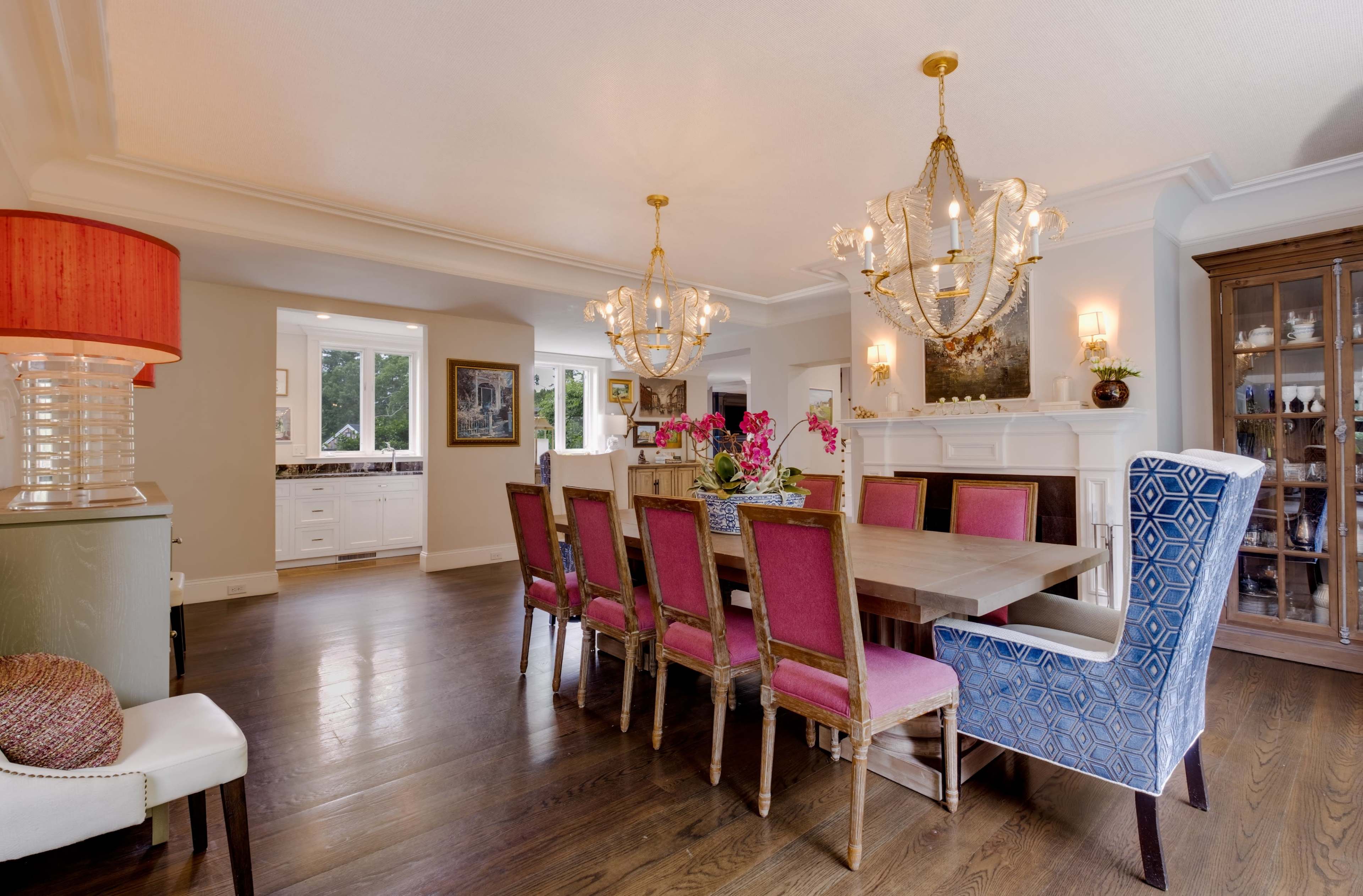 A spacious dining room features a large wooden table surrounded by pink-upholstered chairs, two chandeliers, and a large window with natural light.