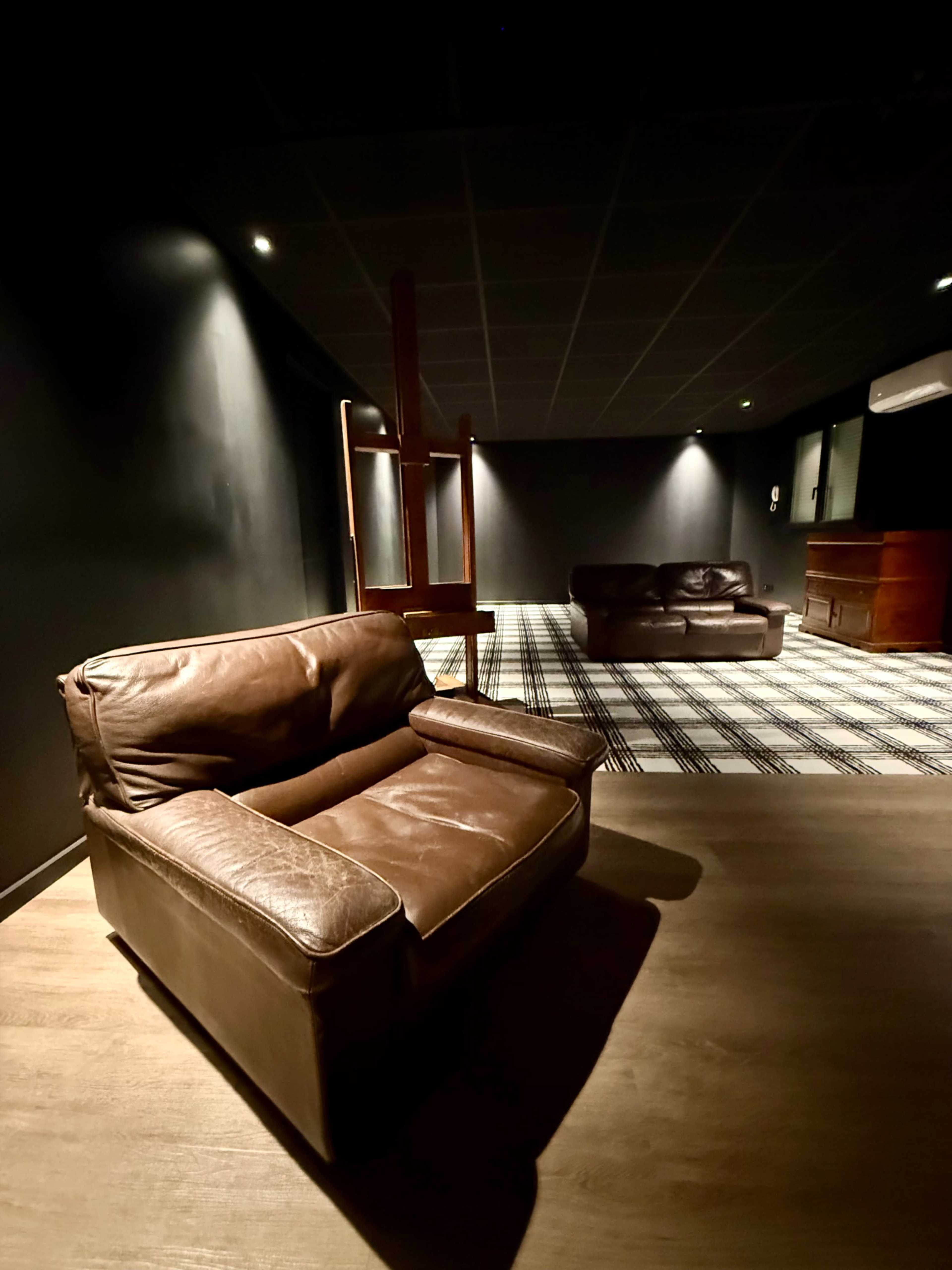 A brown leather armchair is positioned on a patterned floor in a dimly lit room featuring a second couch and an easel in the background.