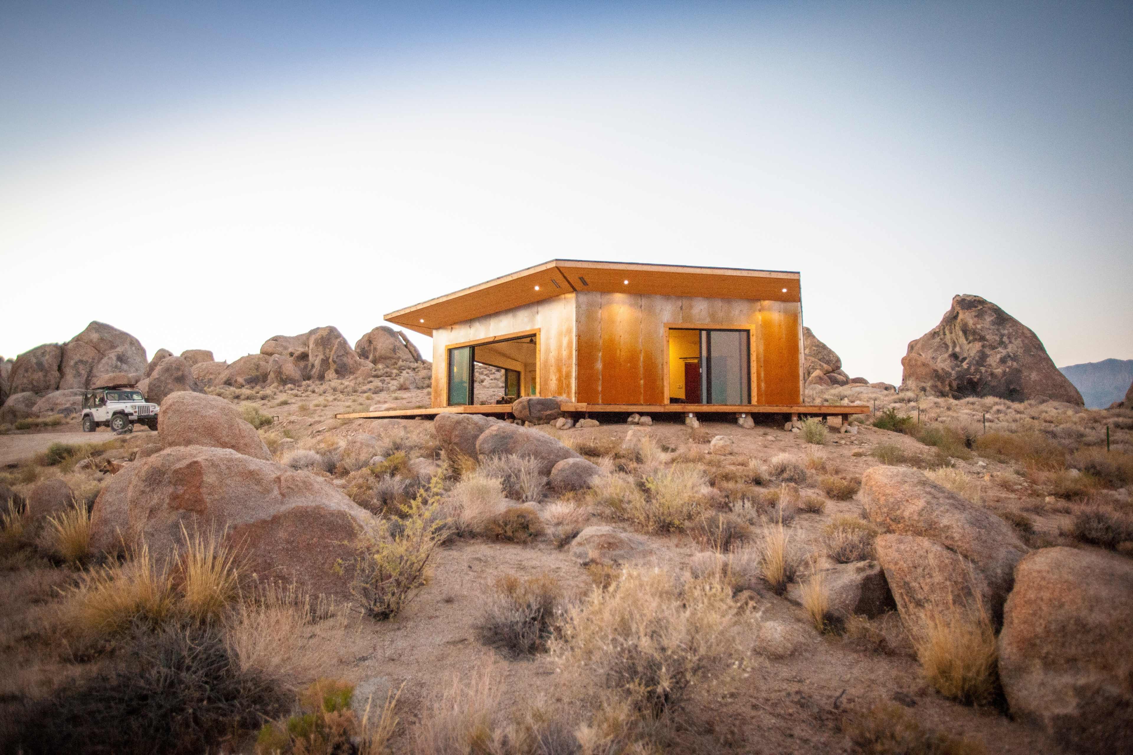 A modern, single-story house with a metal exterior is situated among large boulders in a desert landscape.